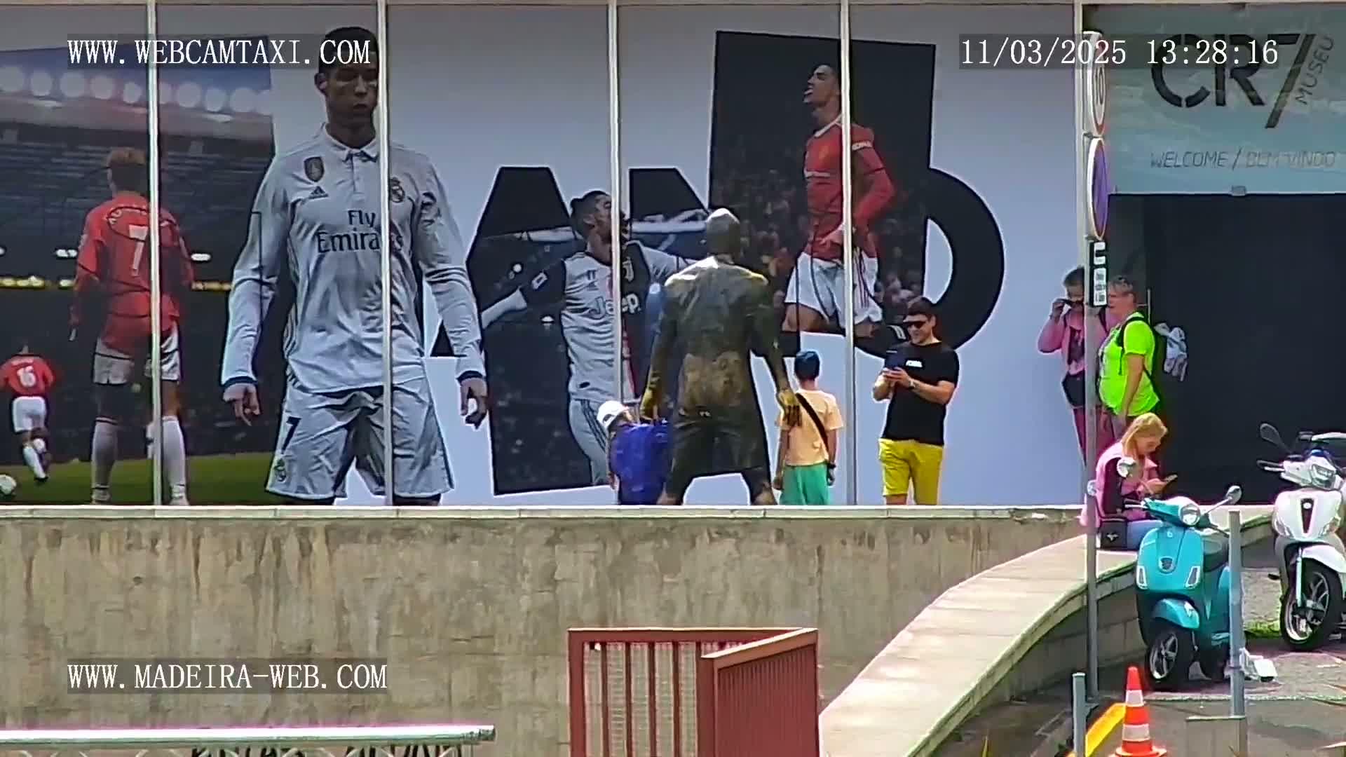 A large building displays images of soccer players, while several people stand and sit in front of it on a sunny day, with a few scooters parked nearby.