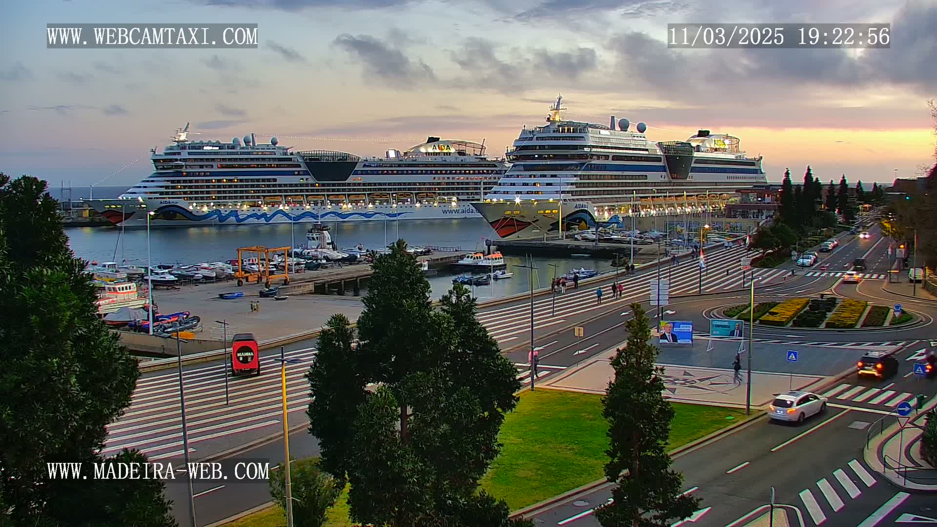 Two large cruise ships are docked at a harbor during a twilight sky, with several cars driving on a nearby road.