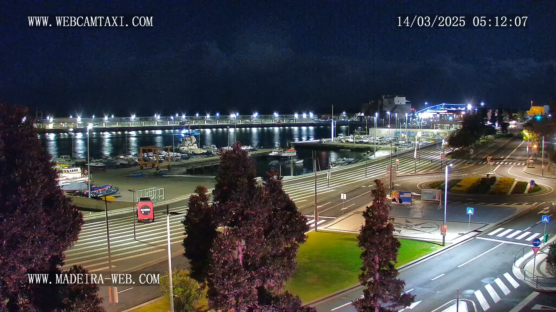 A nighttime view of a harbor filled with boats, adjacent to a roadway with well-lit traffic circles and landscaping.