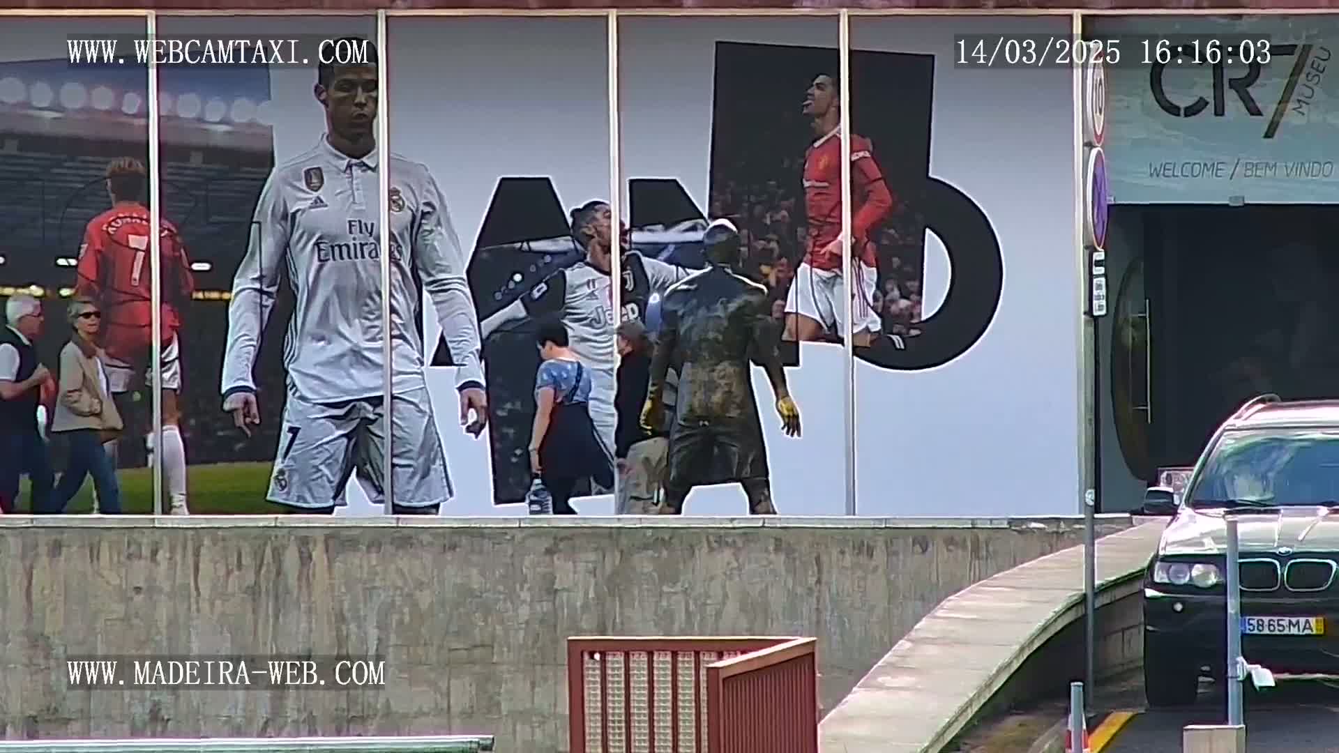 A building exterior displays large images of Cristiano Ronaldo in various soccer uniforms, with several people standing in front of it and a dark-colored SUV parked nearby on a mostly sunny day.