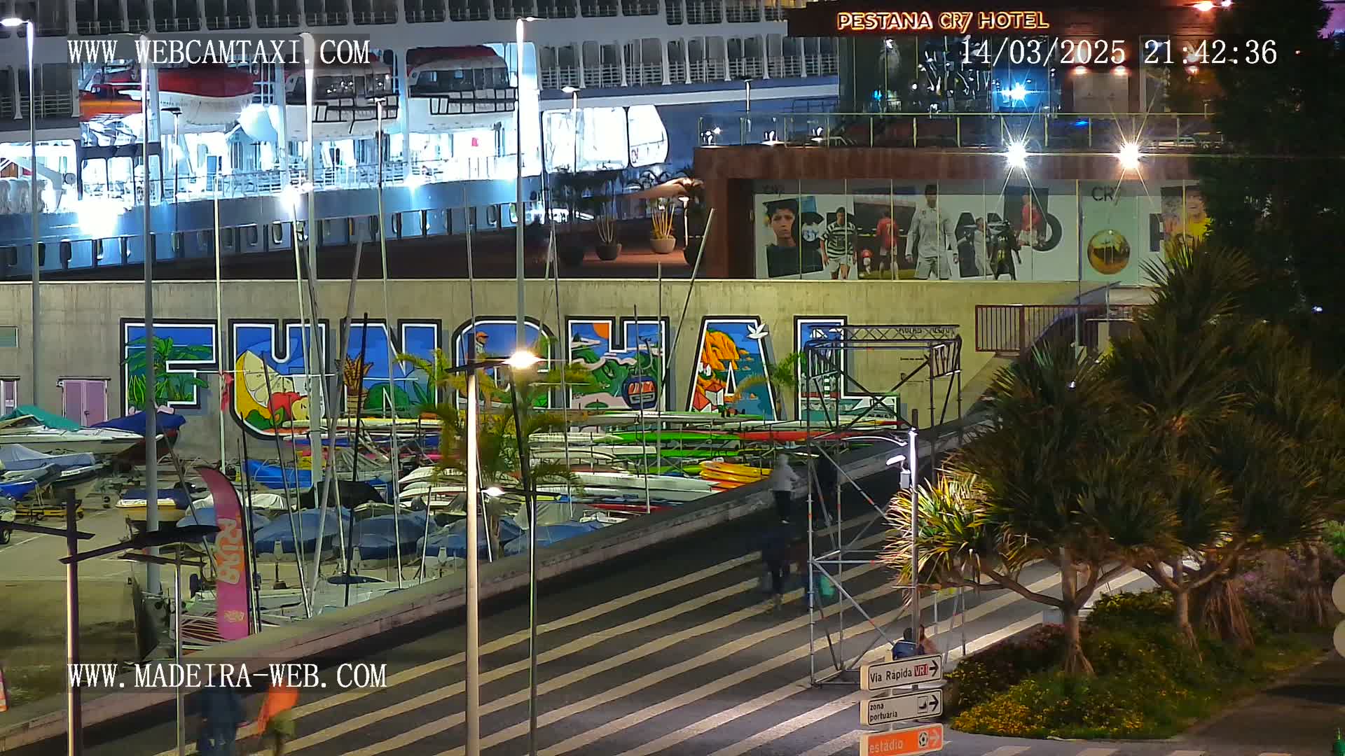 At night, a large cruise ship is docked near a harbor with numerous small boats and kayaks, and a brightly colored mural depicting the name of a location.