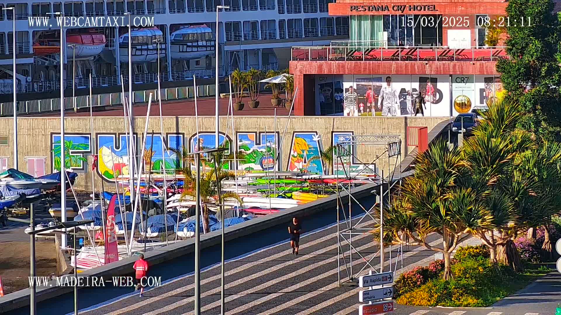 A sunny day shows a harbor with numerous boats and kayaks docked, a large mural depicting the name of a city, a cruise ship in the background, and a person jogging along a walkway.