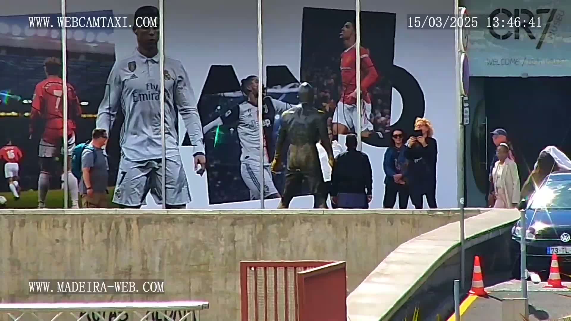 A large billboard depicting soccer players is visible behind a low wall, with several people standing in front of it on a sunny day.
