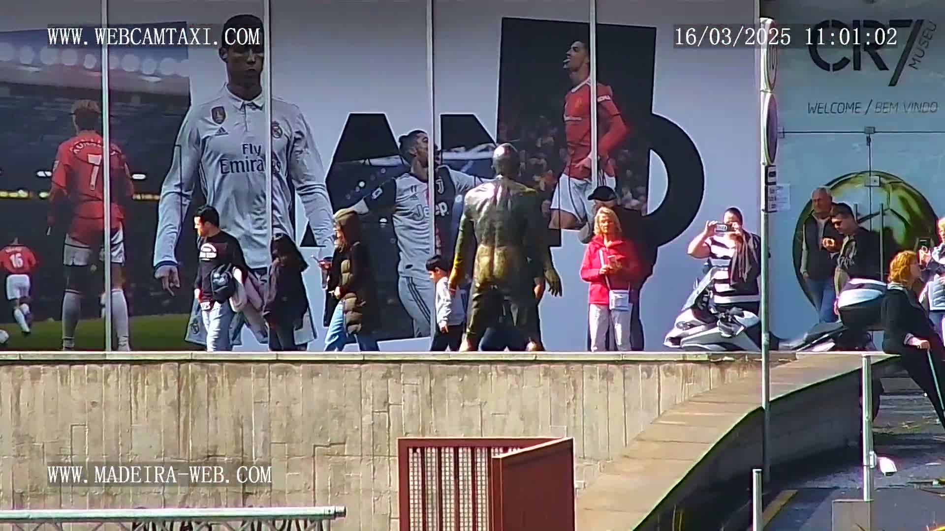 A bronze statue stands in front of a large billboard displaying images of soccer players, while several people stand and walk around it on a sunny day.