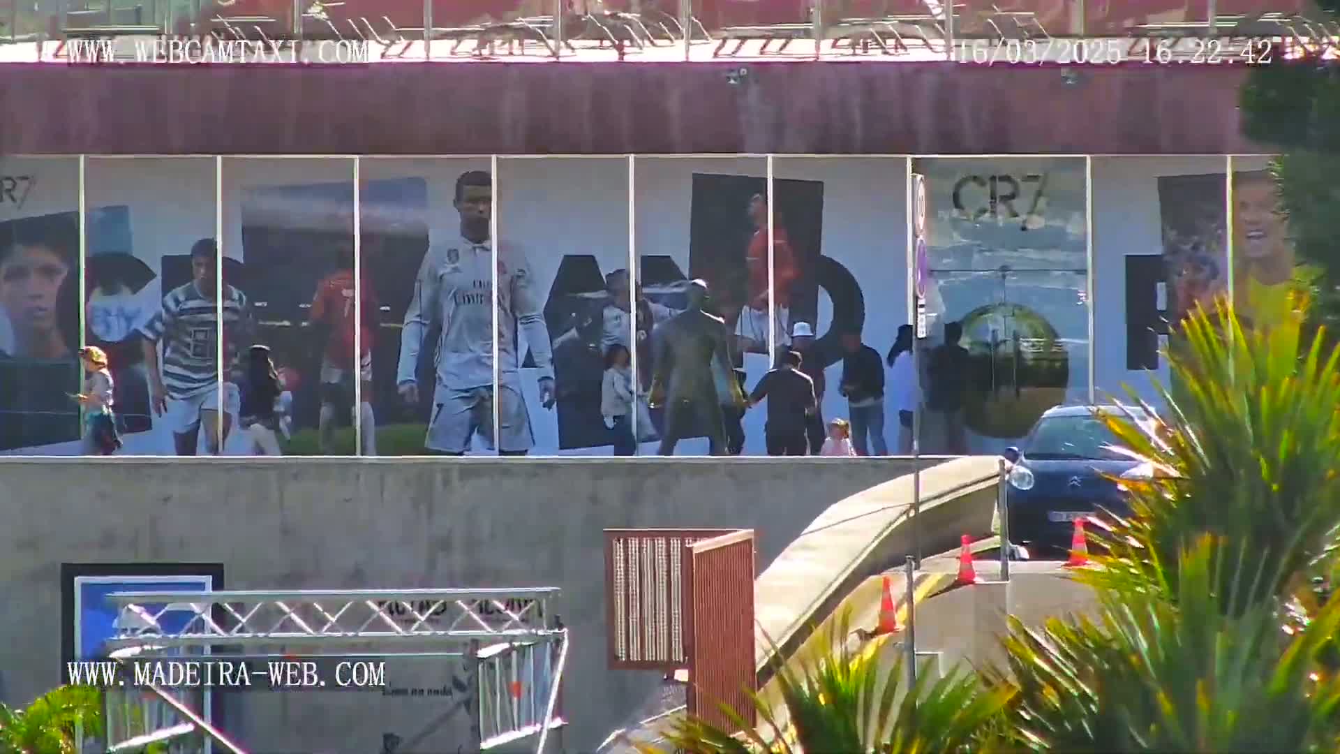 A large building displays images of Cristiano Ronaldo, while people stand in front of it on a sunny day; a car is parked nearby.