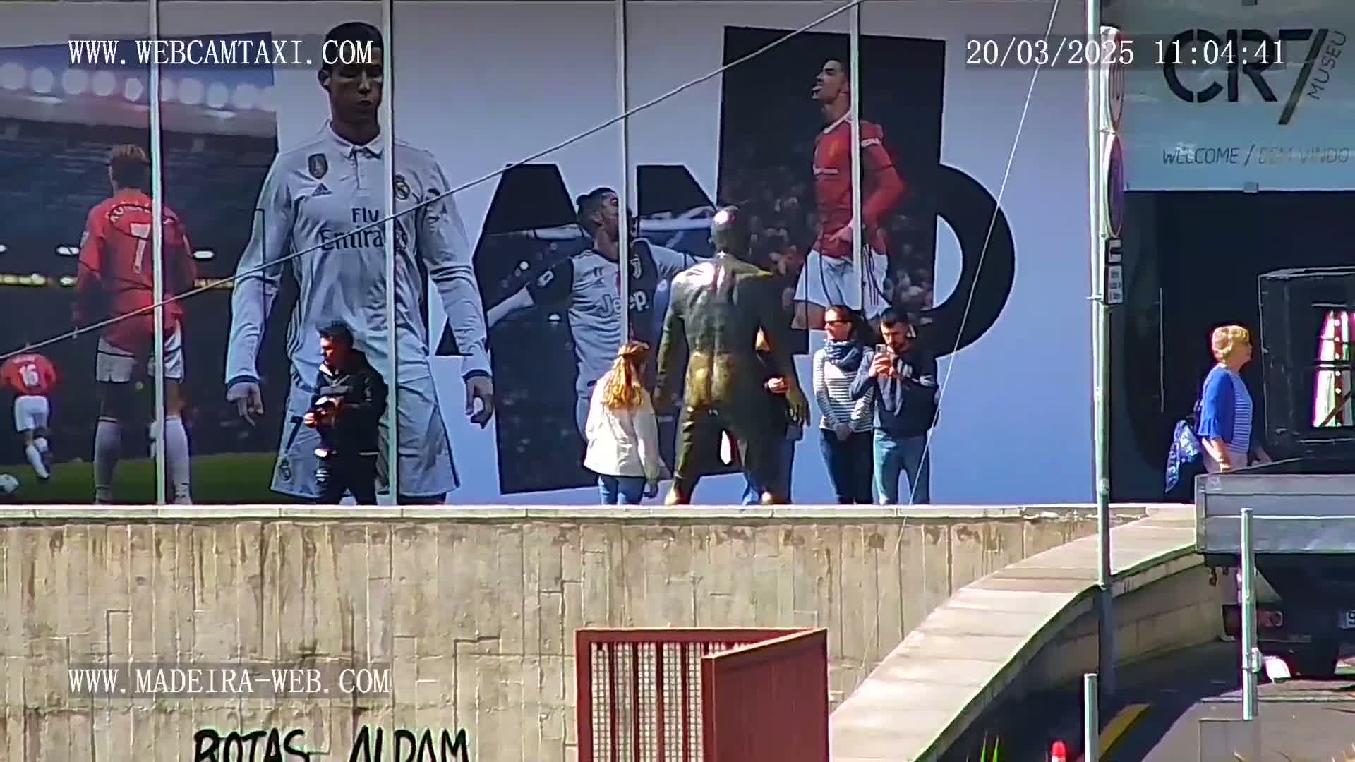 A large billboard depicting soccer players is visible in the sunny daylight, with several people standing in front of it and others walking by on a sidewalk.