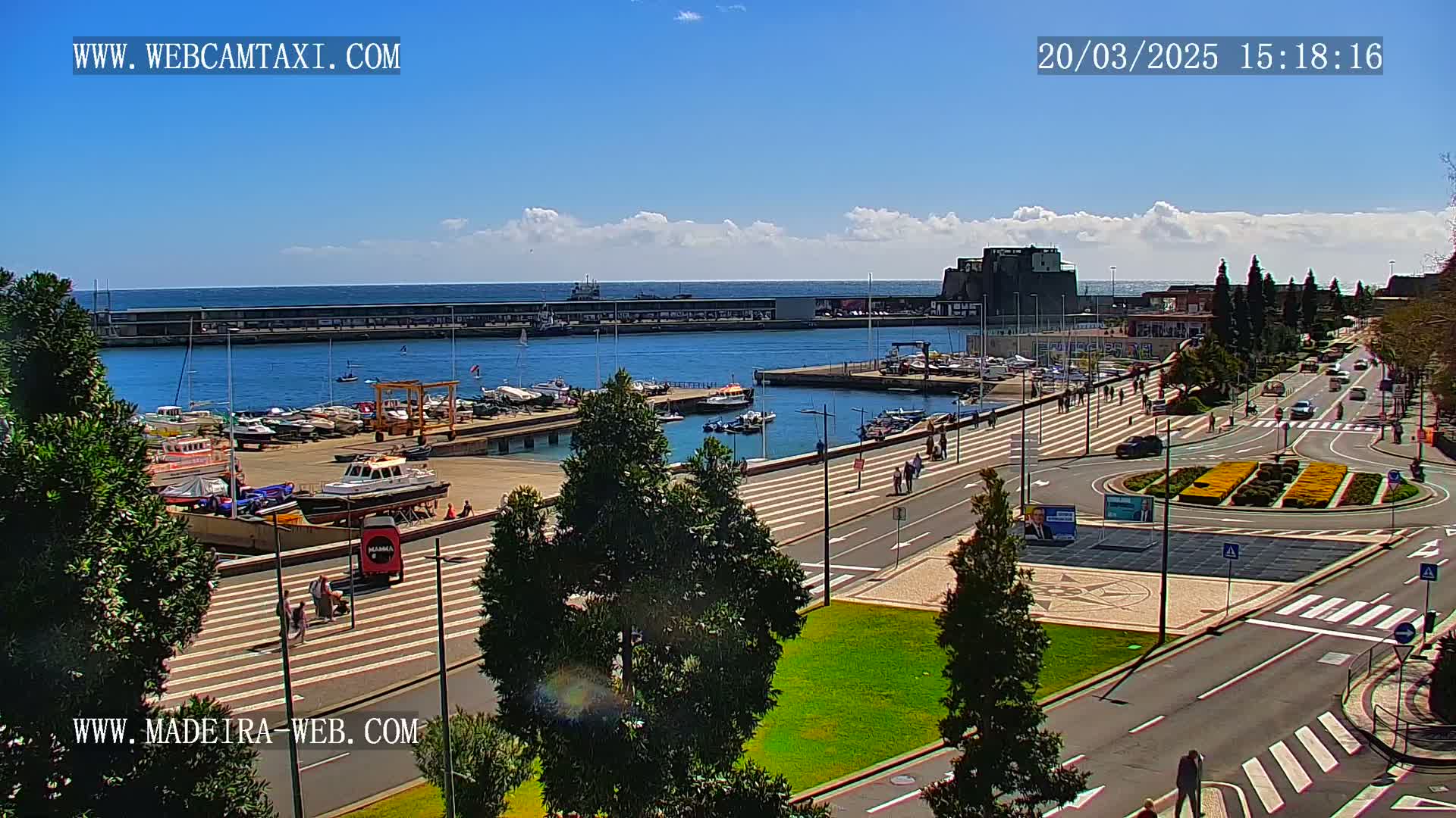 A harbor filled with boats is seen from an elevated vantage point on a sunny day, with a paved waterfront area featuring pedestrians and roadways leading to a roundabout.