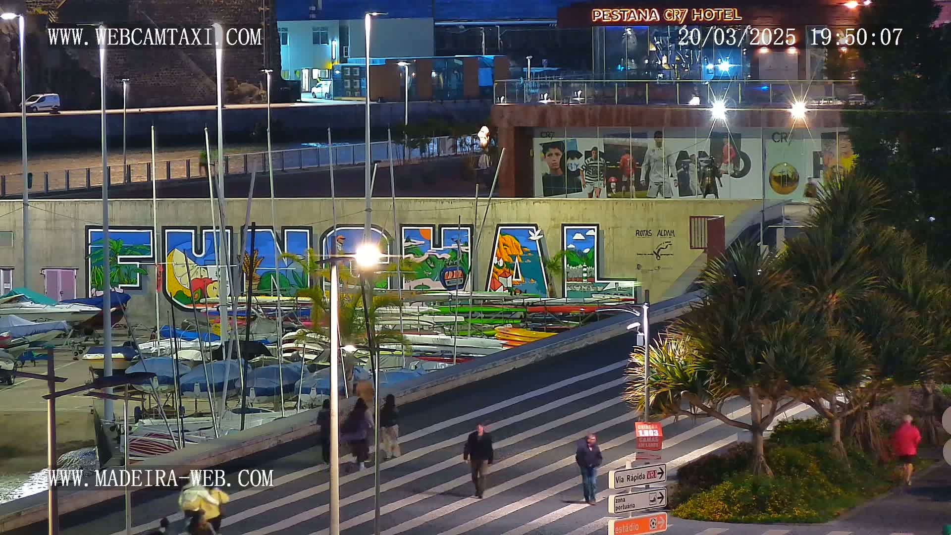 A marina at night, with numerous boats docked alongside a wall featuring a large mural of the word "Funchal," is illuminated by streetlights under a clear, dark sky.
