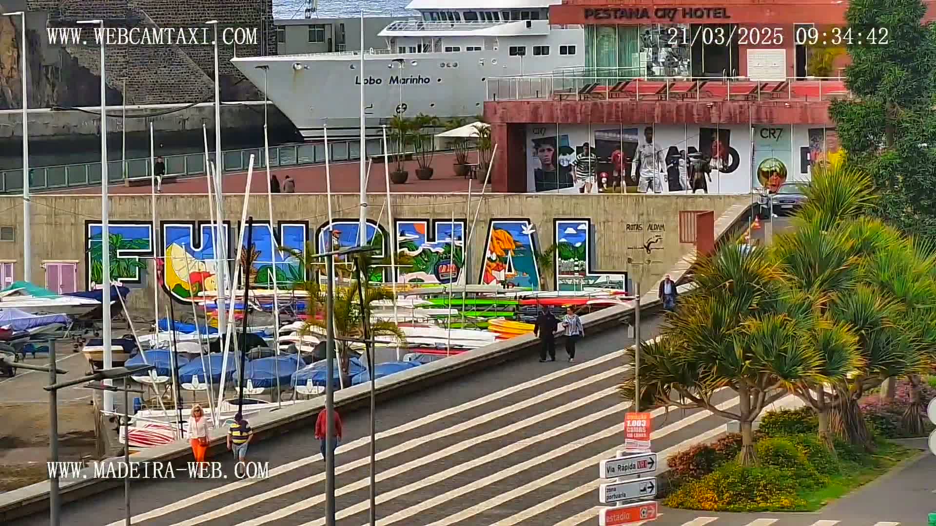 A harbor scene on a sunny day shows a large mural of the word "Funchal," numerous boats docked, and people walking along a paved walkway next to a building with advertisements.