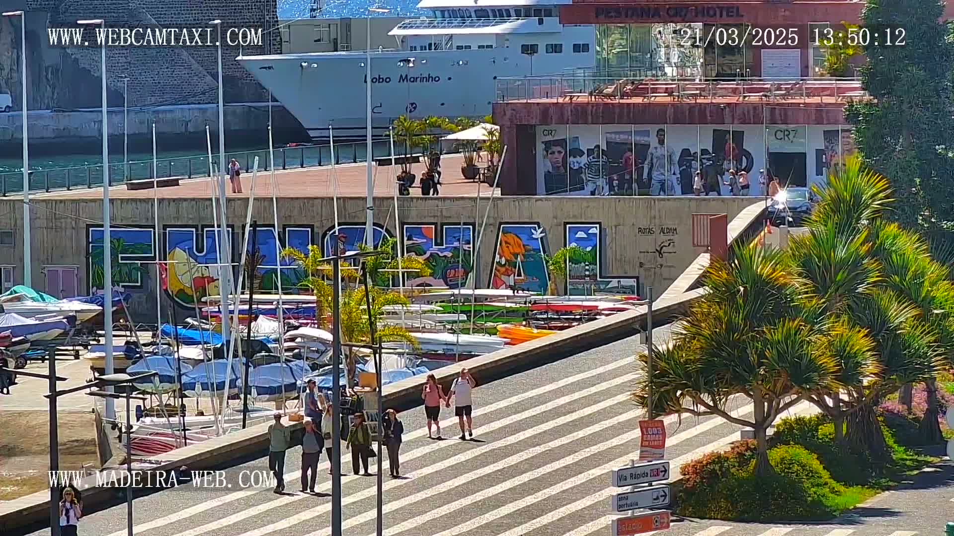 A sunny harbor scene features a large mural, numerous boats, a cruise ship, and pedestrians walking along a striped walkway.