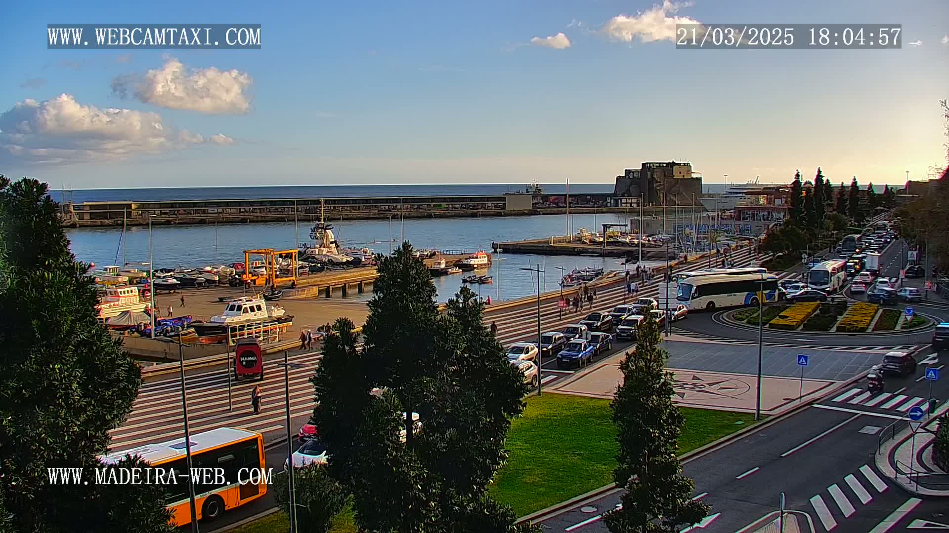 A harbor with numerous boats and cars parked along a waterfront promenade under a partly cloudy sky.