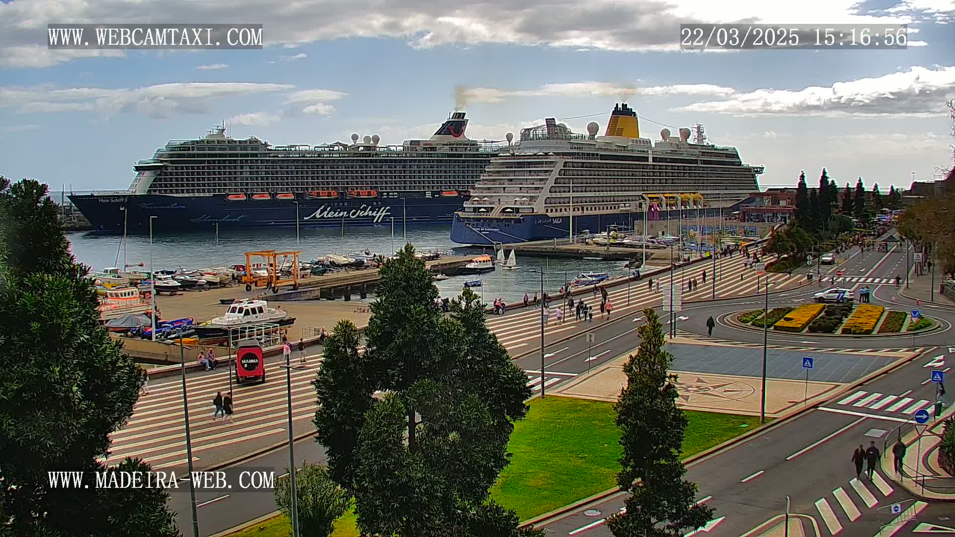 Two large cruise ships are docked at a harbor, with pedestrians walking along a paved area next to the water under partly cloudy skies.