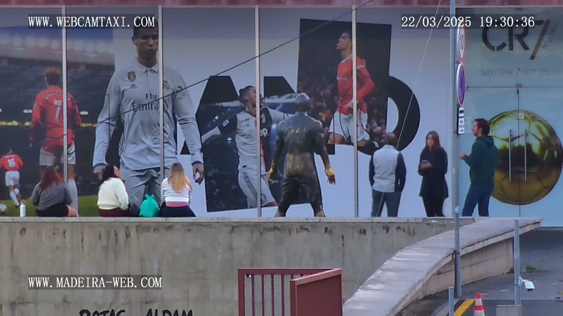 A large mural of Cristiano Ronaldo in different soccer uniforms is displayed on a building, with several people standing in front of it on a somewhat overcast day.