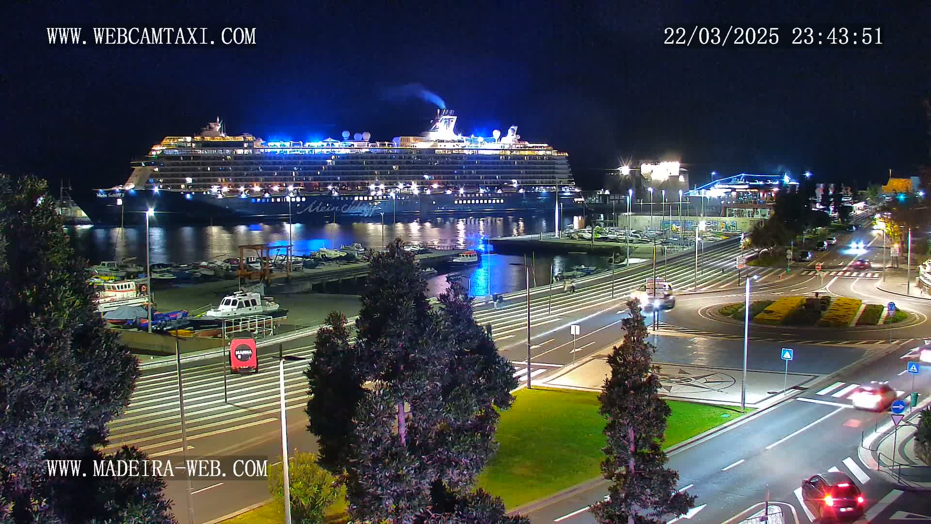 A large cruise ship is docked at a harbor at night, with cars driving on a nearby road.