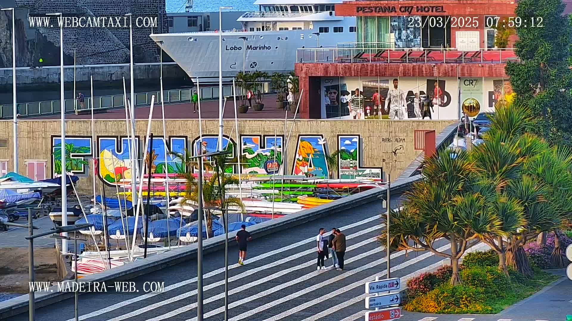 A colorful mural of "Funchal" is visible behind a harbor filled with boats, with a person jogging along a walkway overlooking the scene on a sunny day.