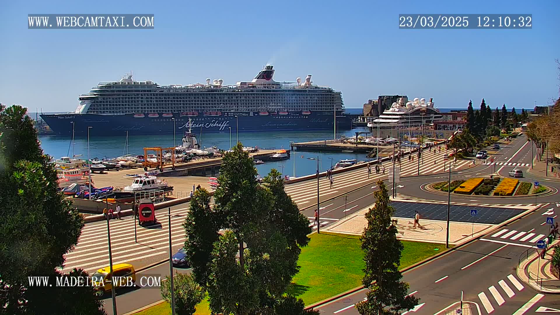 A large cruise ship is docked at a harbor on a sunny day, with smaller boats and a paved waterfront area visible, including pedestrians and roadways.