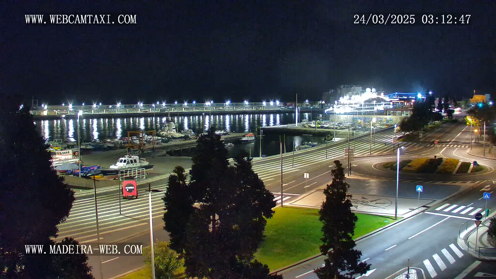 A nighttime view of a harbor filled with boats, alongside a roadway with minimal traffic under clear skies.