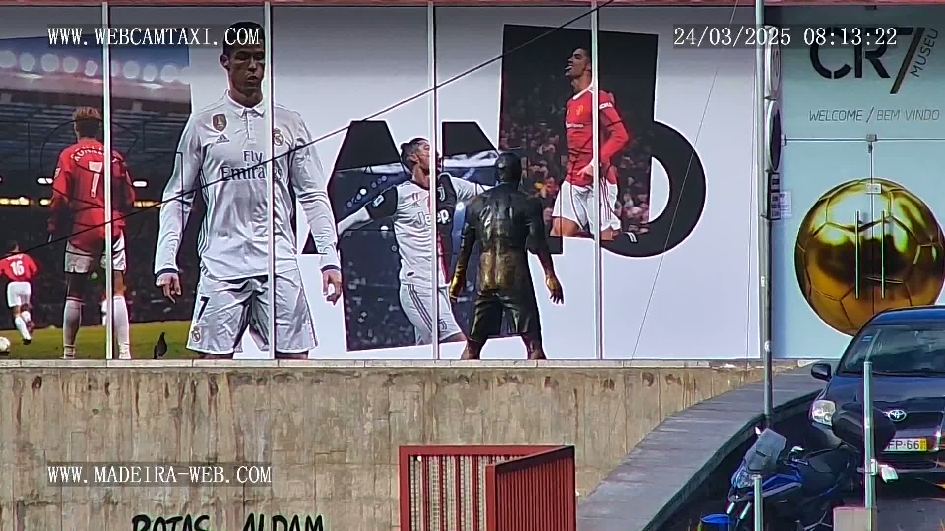 A large billboard displays images of Cristiano Ronaldo in various soccer uniforms, a golden soccer ball, and a dark-colored statue; a sunny day is visible in the background.