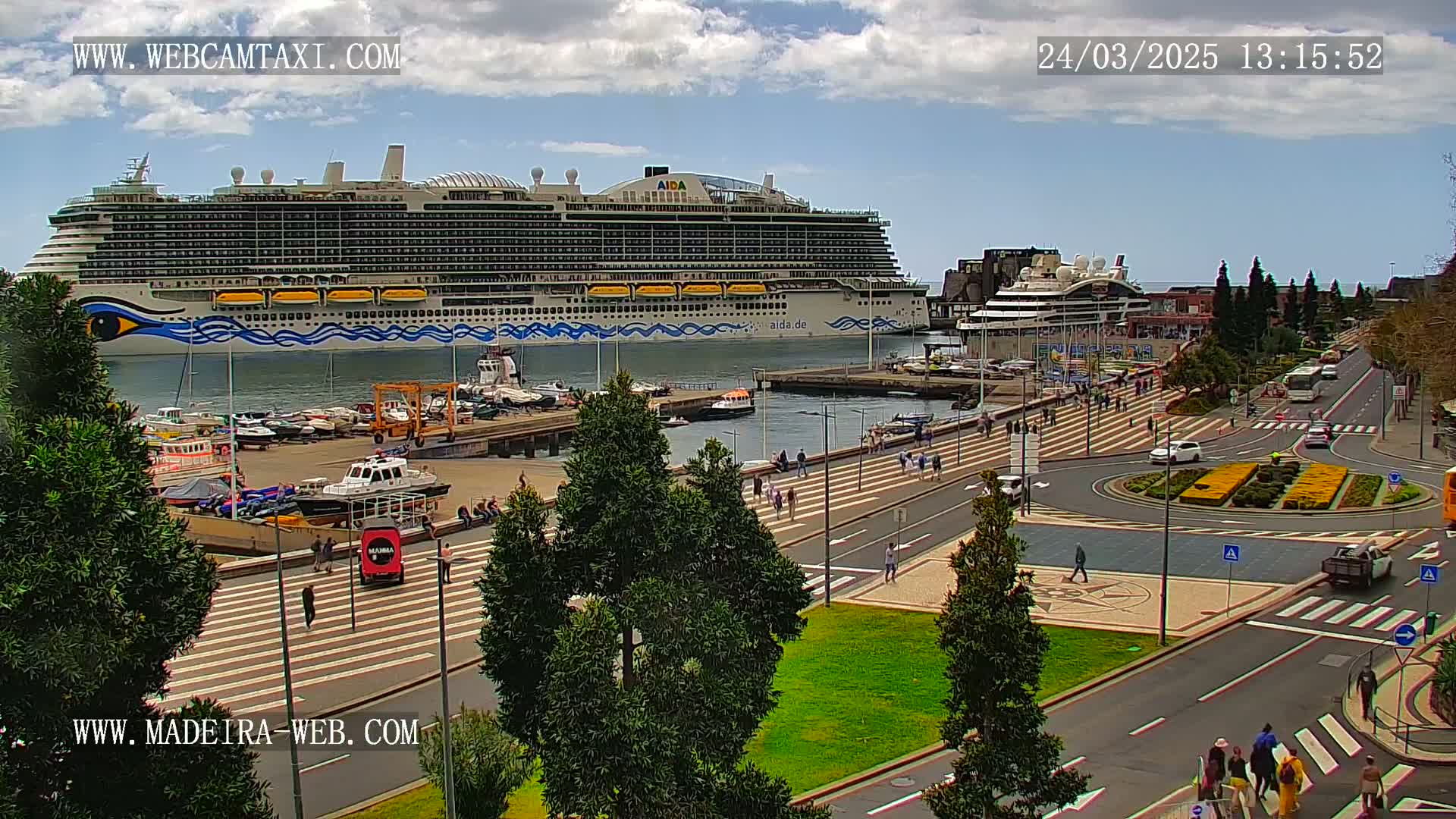 A large cruise ship is docked at a port, with pedestrians walking along a paved waterfront area under partly cloudy skies.