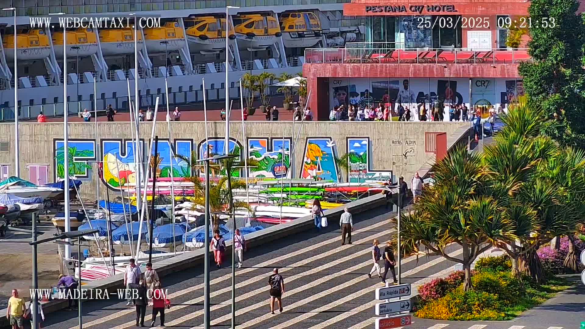 A harbor scene on a sunny day features a large mural of the word "Funchal," numerous boats docked, pedestrians walking along a striped walkway, and a cruise ship in the background.