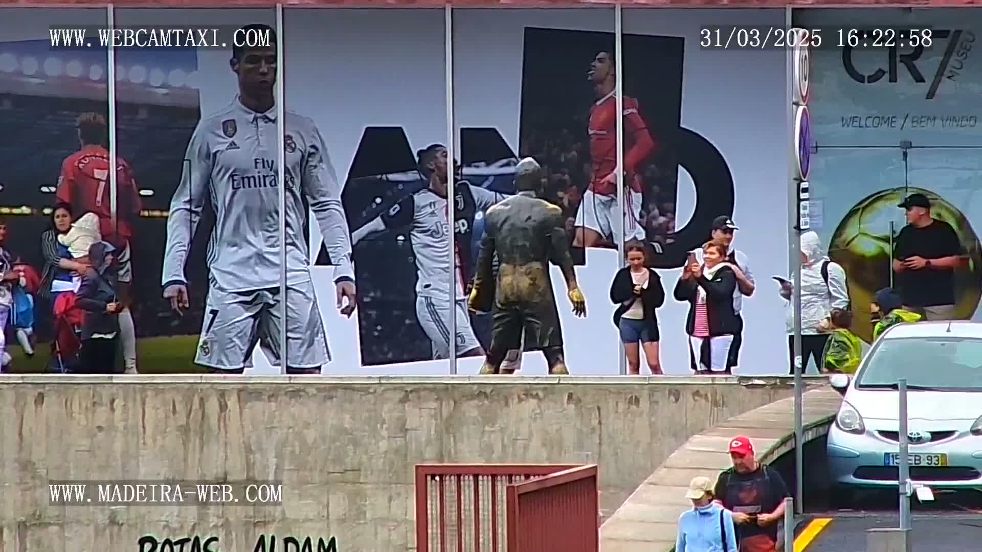 A large billboard displaying images of Cristiano Ronaldo and other soccer players is visible behind a group of people and cars on an overcast day.