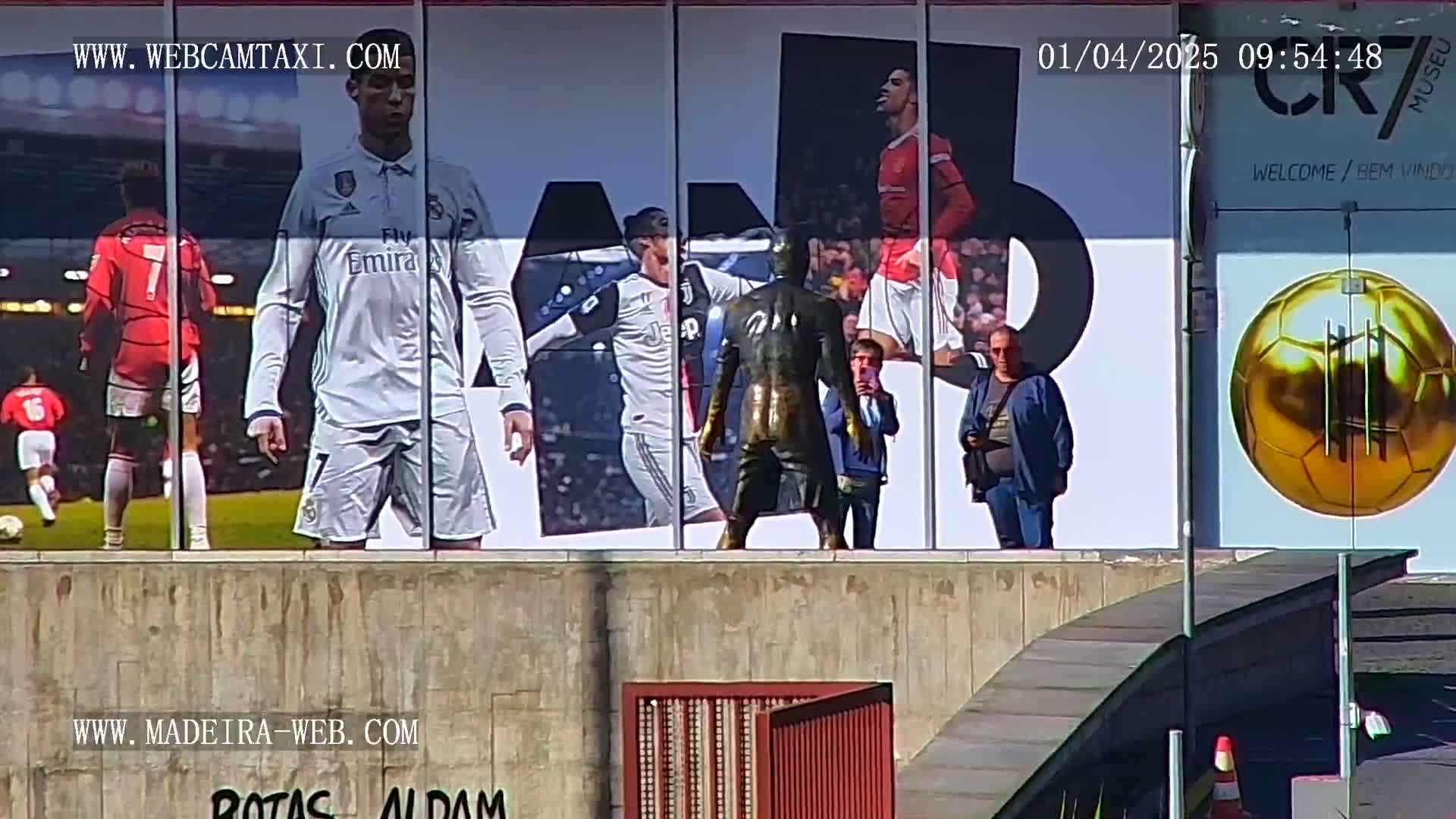 A large billboard displaying images of soccer players is mounted on a building exterior on a sunny day, with several people standing in front of it.