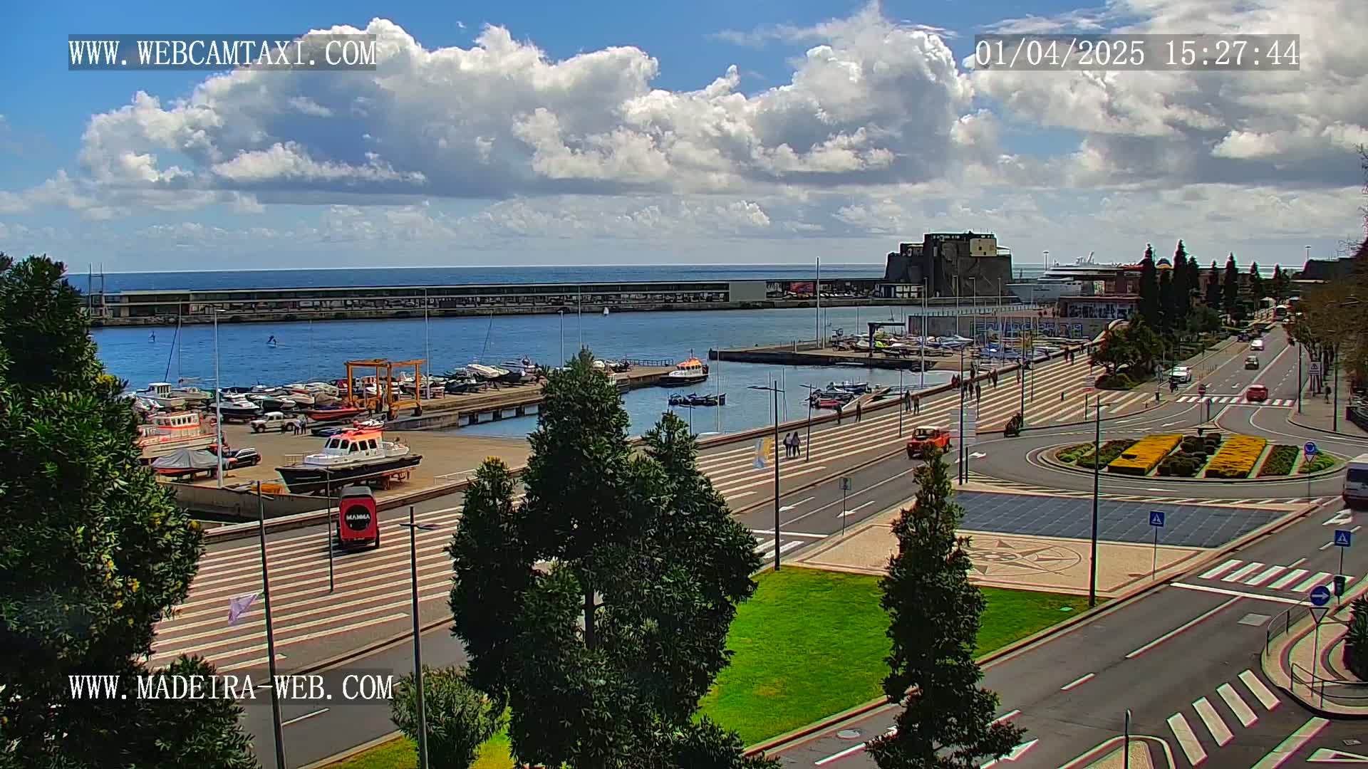 A harbor filled with boats is visible under a partly cloudy sky, with a paved area and roadway leading away from the waterfront.