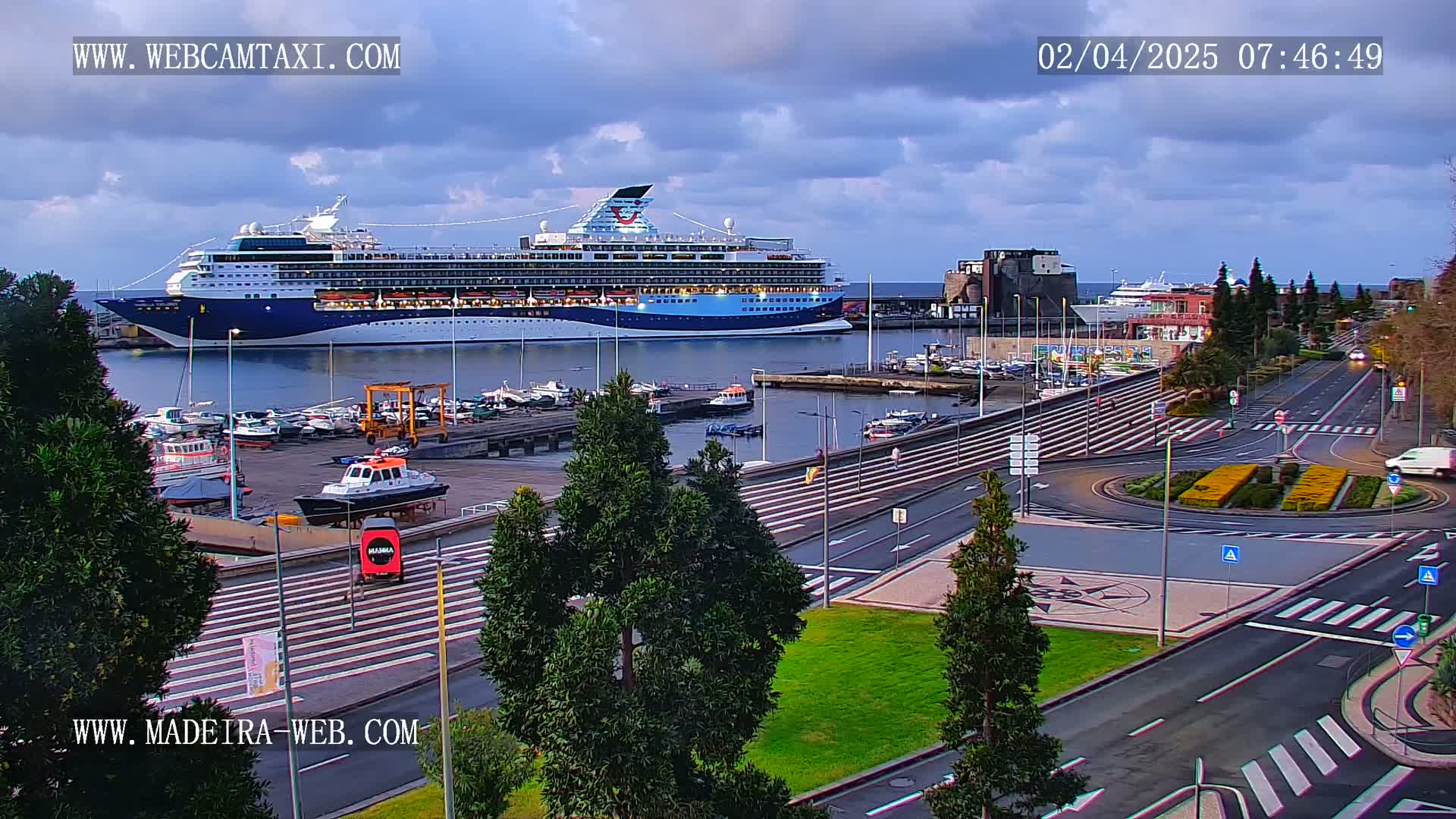 A large cruise ship is docked at a harbor, with smaller boats in the marina, under a partly cloudy sky.