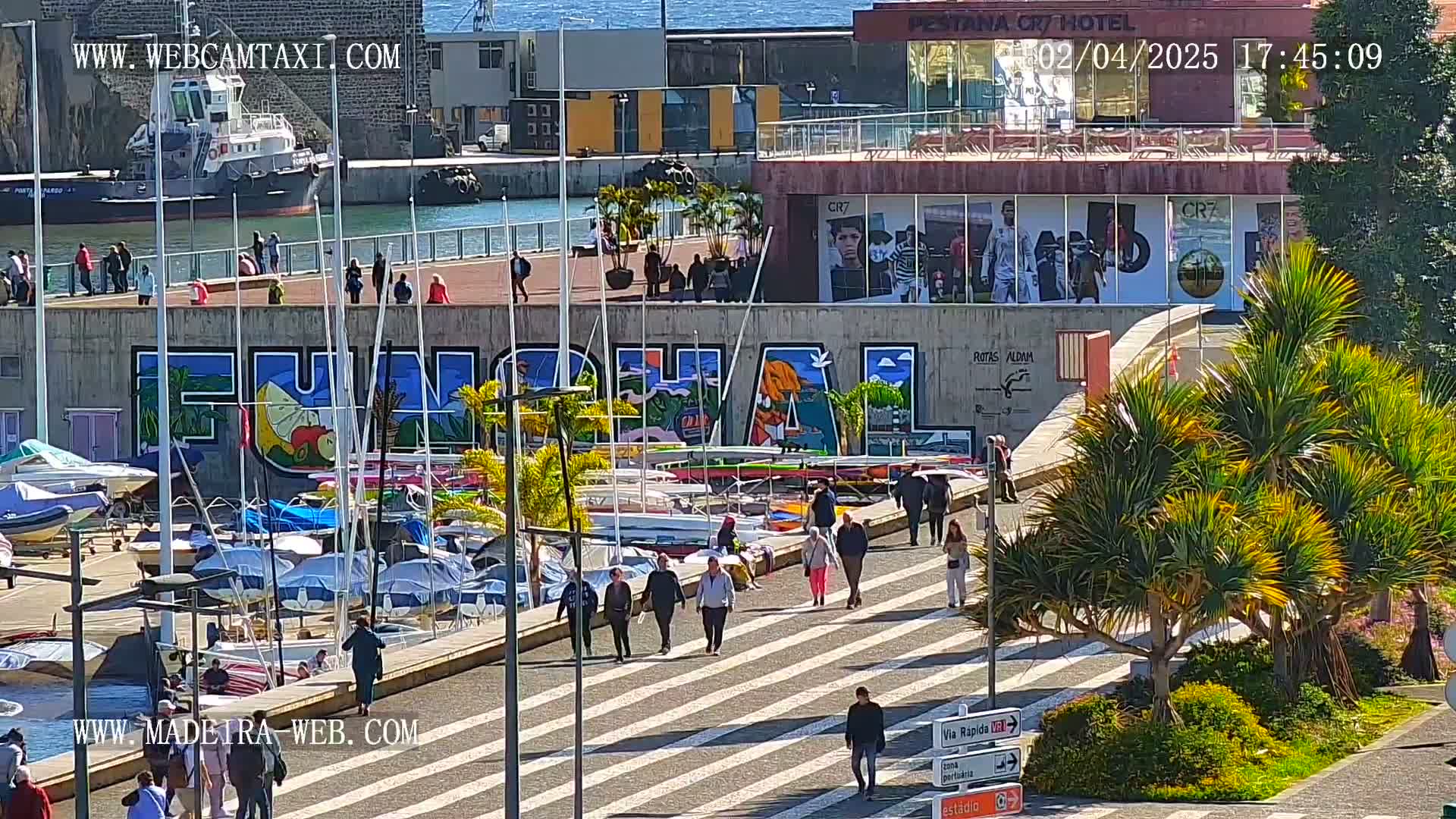 A sunny harbor scene shows numerous boats docked, people walking along a paved walkway, and a large mural on a wall.
