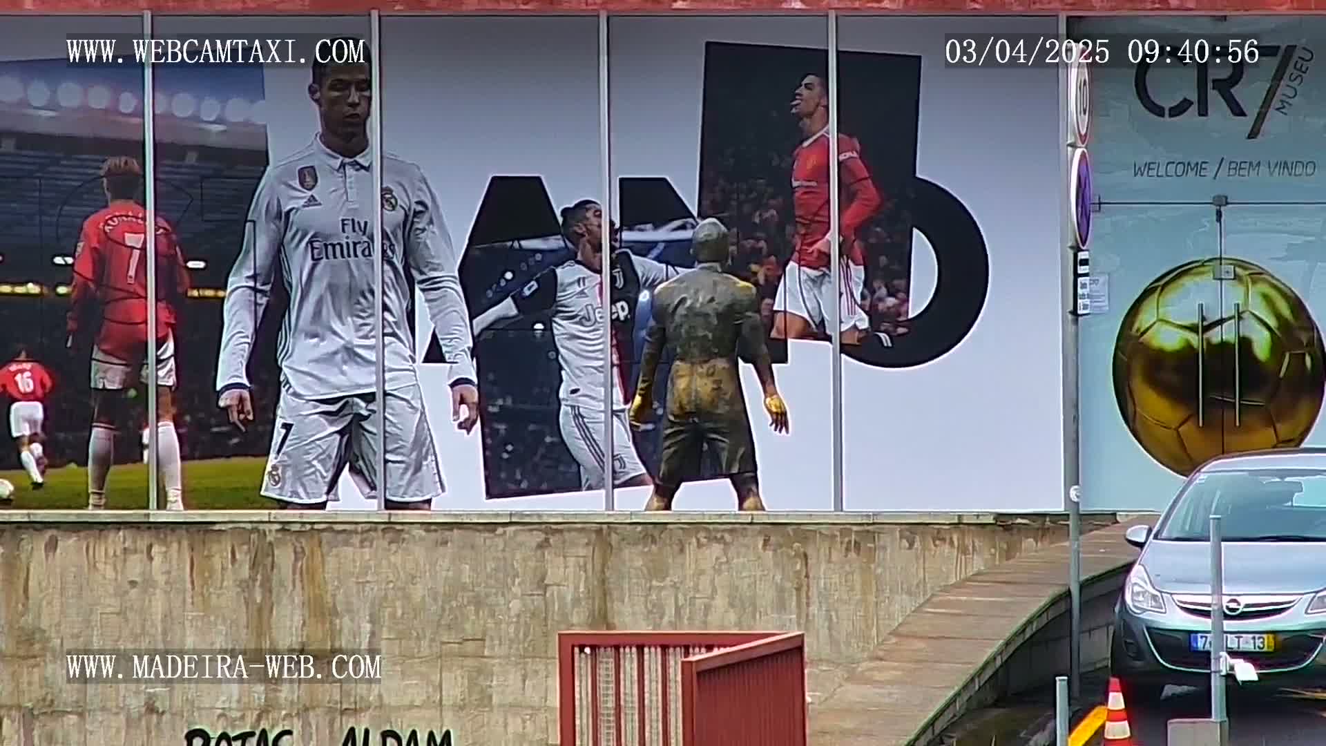 A large building displays a mural of several soccer players, a statue of a soccer player, and a large golden soccer ball; a silver car is parked in front of it on an overcast day.