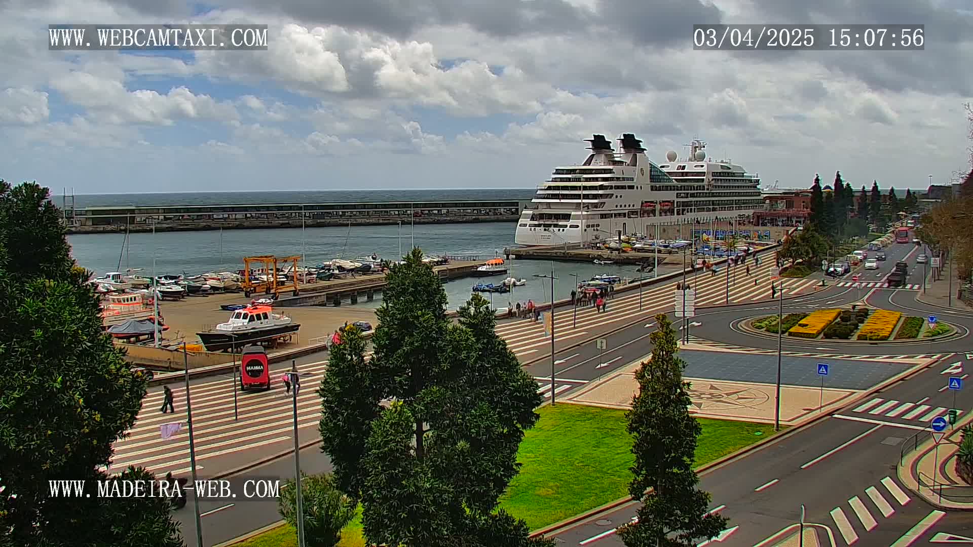 A large cruise ship is docked at a harbor, with a partly cloudy sky above and pedestrians walking along a paved waterfront area next to a roadway.