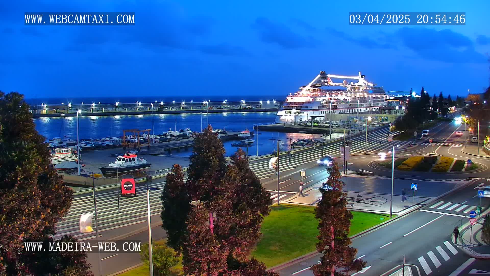A large cruise ship is docked at a harbor at twilight, with cars driving on roads and smaller boats in the marina.