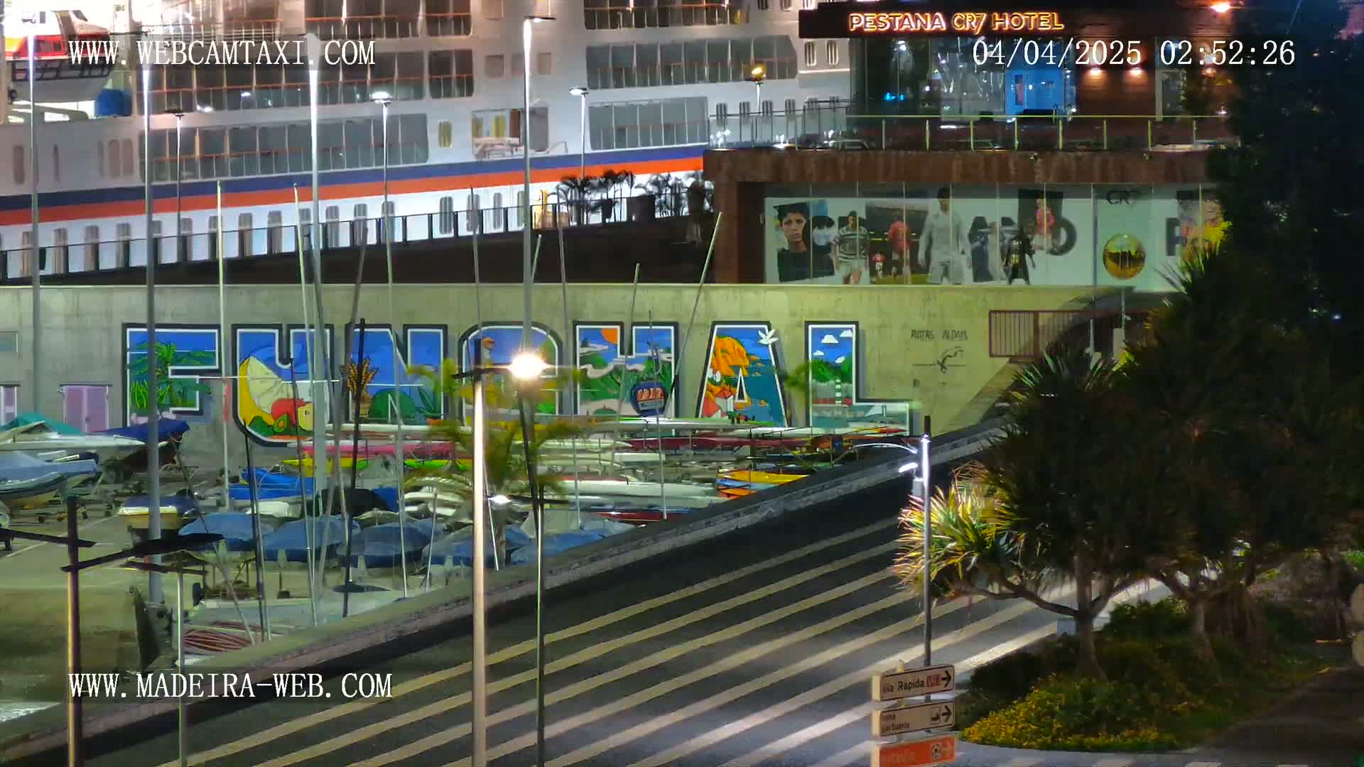 At night, a large cruise ship is docked near a marina filled with small boats, and a mural depicting the word "Funchal" is visible on a wall beside a hotel.