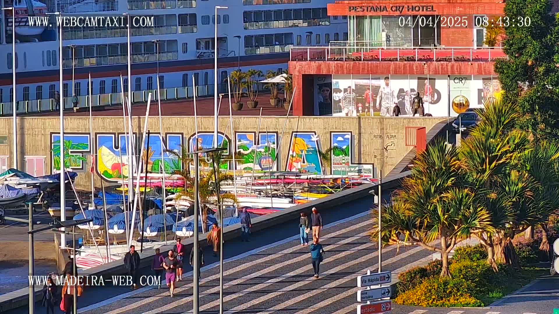 A sunny harbor scene shows a large mural of "Funchal" on a building behind a marina filled with sailboats and small motorboats, with pedestrians walking along a paved walkway in front of the building.