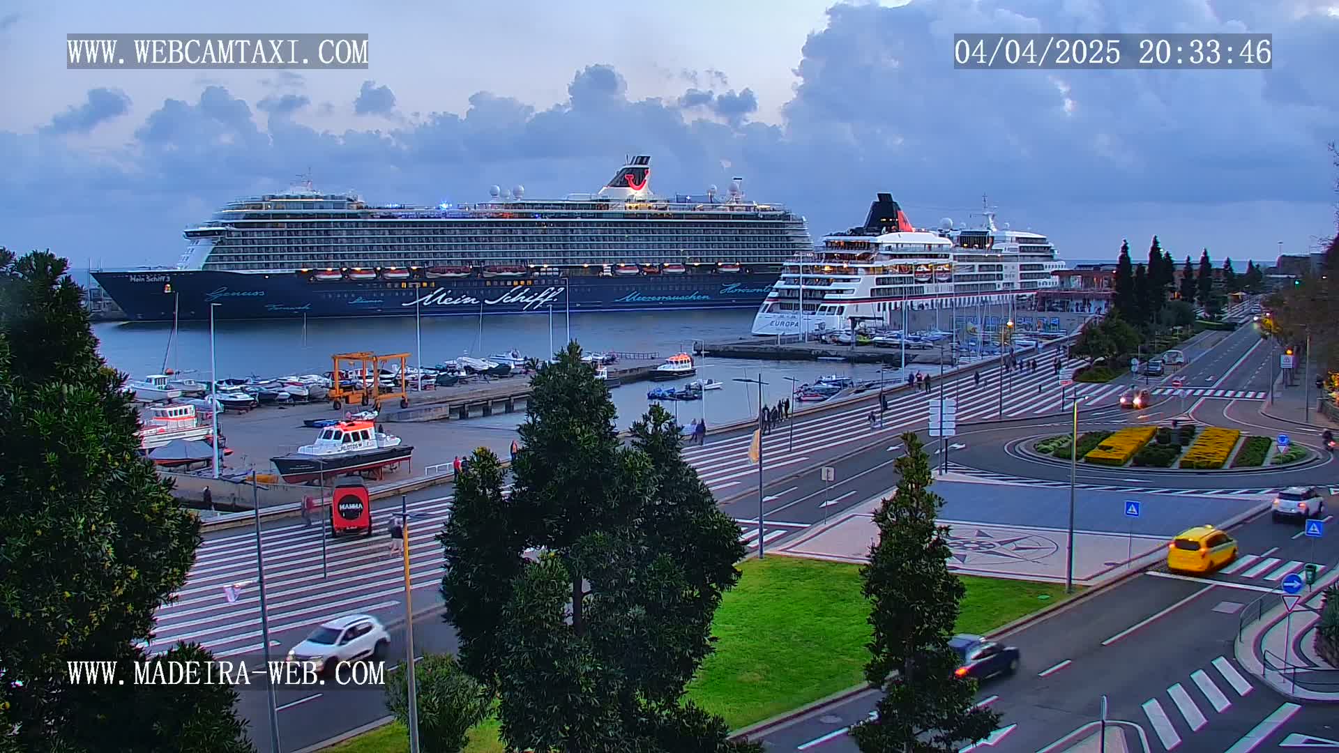 Two large cruise ships are docked at a port, with several smaller boats in the harbor, under an overcast twilight sky; a roadway with pedestrian crossings and several cars is visible in the foreground.