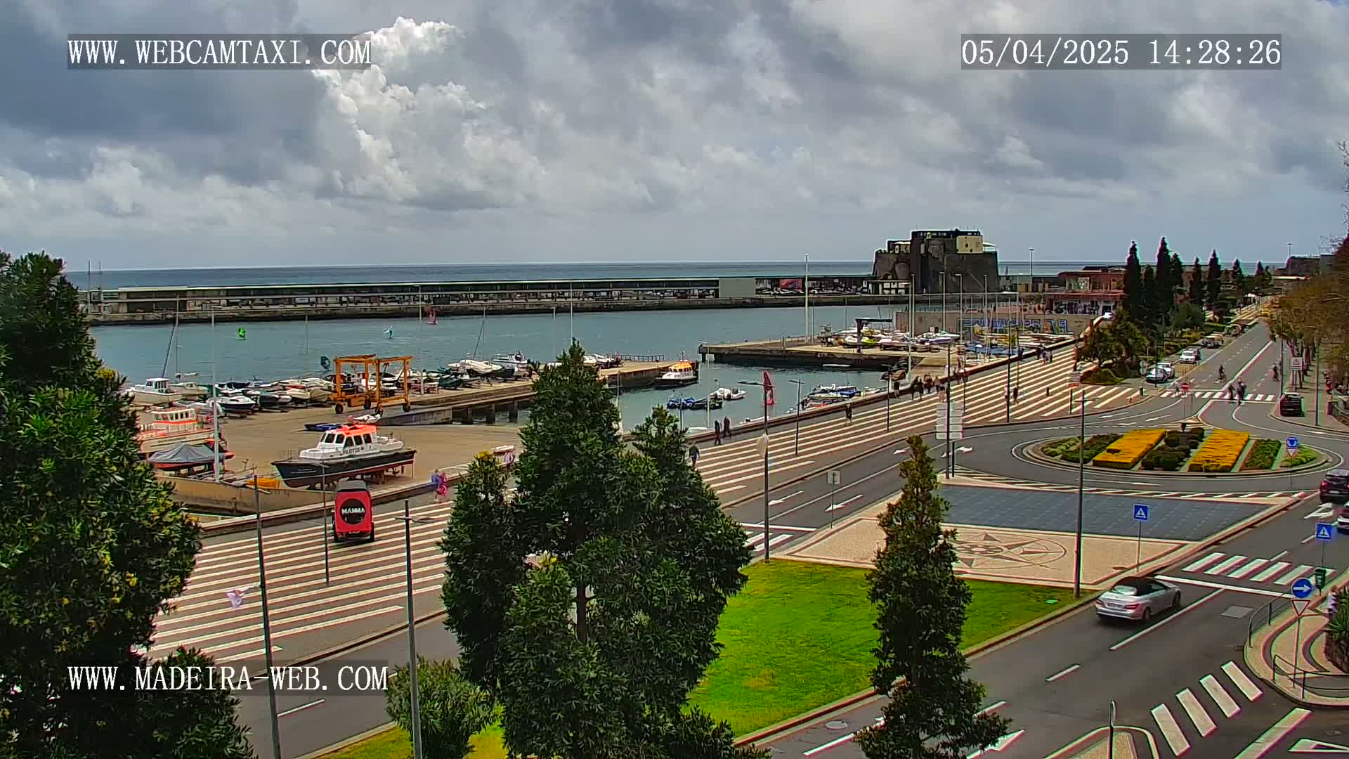 A harbor filled with boats is next to a paved area with trees and a roundabout, all under a partly cloudy sky.