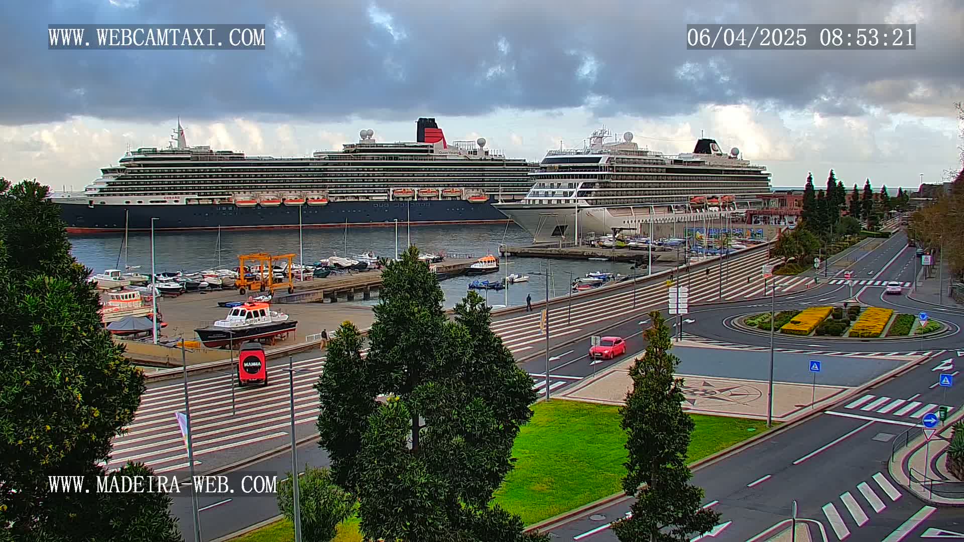 Two large cruise ships are docked at a harbor, with a partly cloudy sky overhead and a paved road with sparse traffic visible in the foreground.