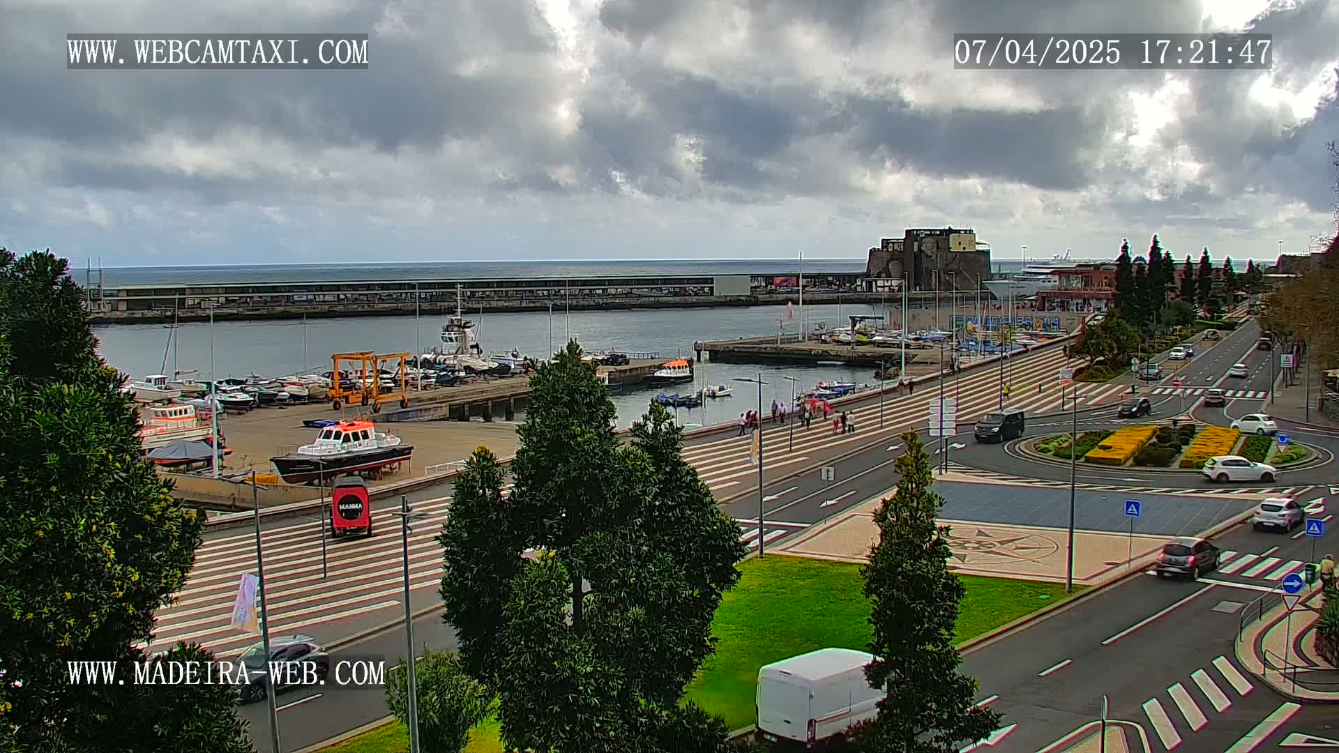 An overcast day shows a harbor filled with docked boats and a roadway with several cars and pedestrians, all situated near a body of water.