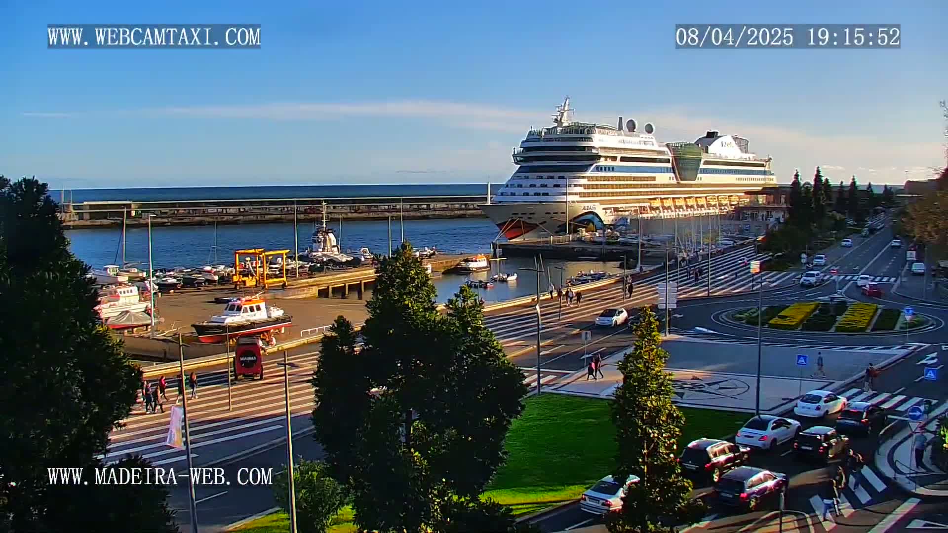 A large cruise ship is docked at a harbor, with cars driving on the roads and pedestrians walking nearby under a clear, sunny sky.