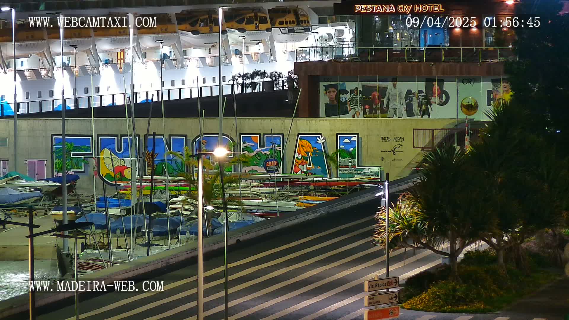 At night, a harbor with numerous boats is shown beneath a large mural that says "Funchal," next to a building with images of soccer players.