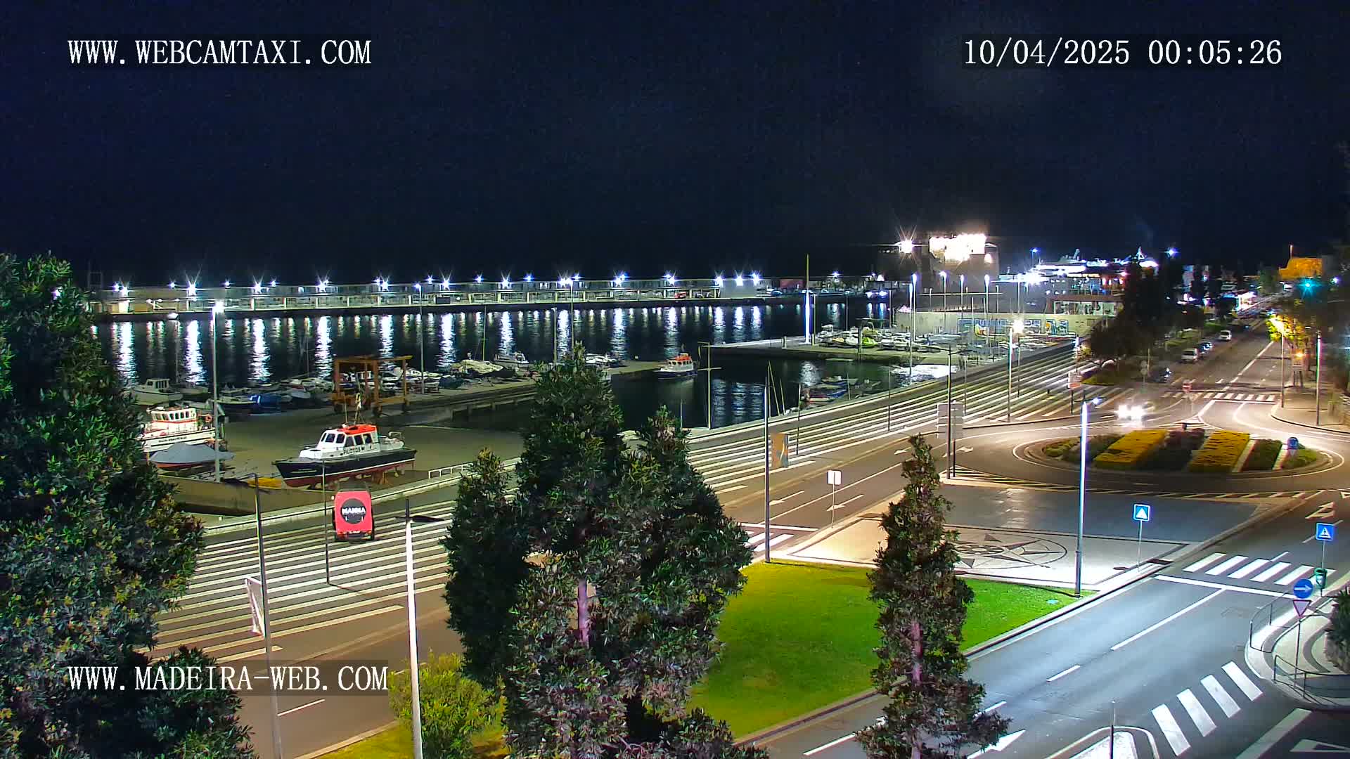 A nighttime view of a harbor filled with boats, alongside a roadway with traffic and greenery under a clear sky.