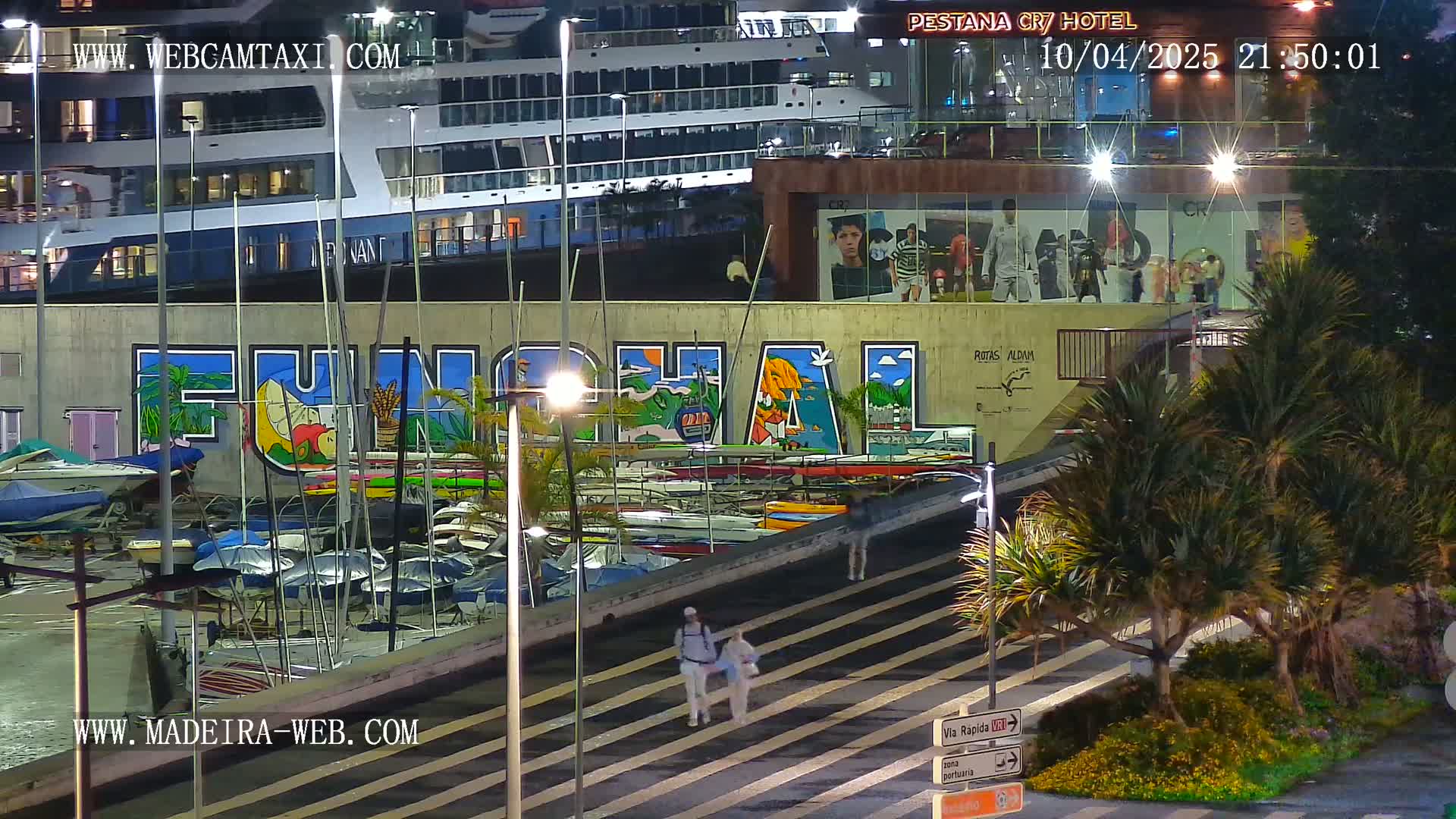 A nighttime harbor scene shows a large cruise ship docked near a wall with a mural of the word "Funchal," numerous small boats in the water, and two people walking along a paved walkway, flanked by palm trees.