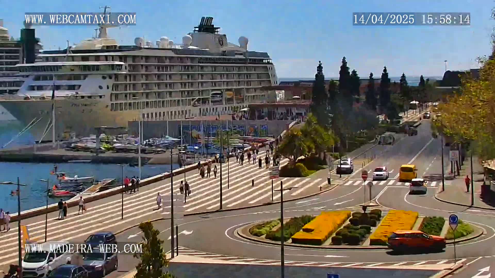A large cruise ship is docked at a harbor on a sunny day, with pedestrians walking along a paved waterfront area and cars driving on a nearby road.