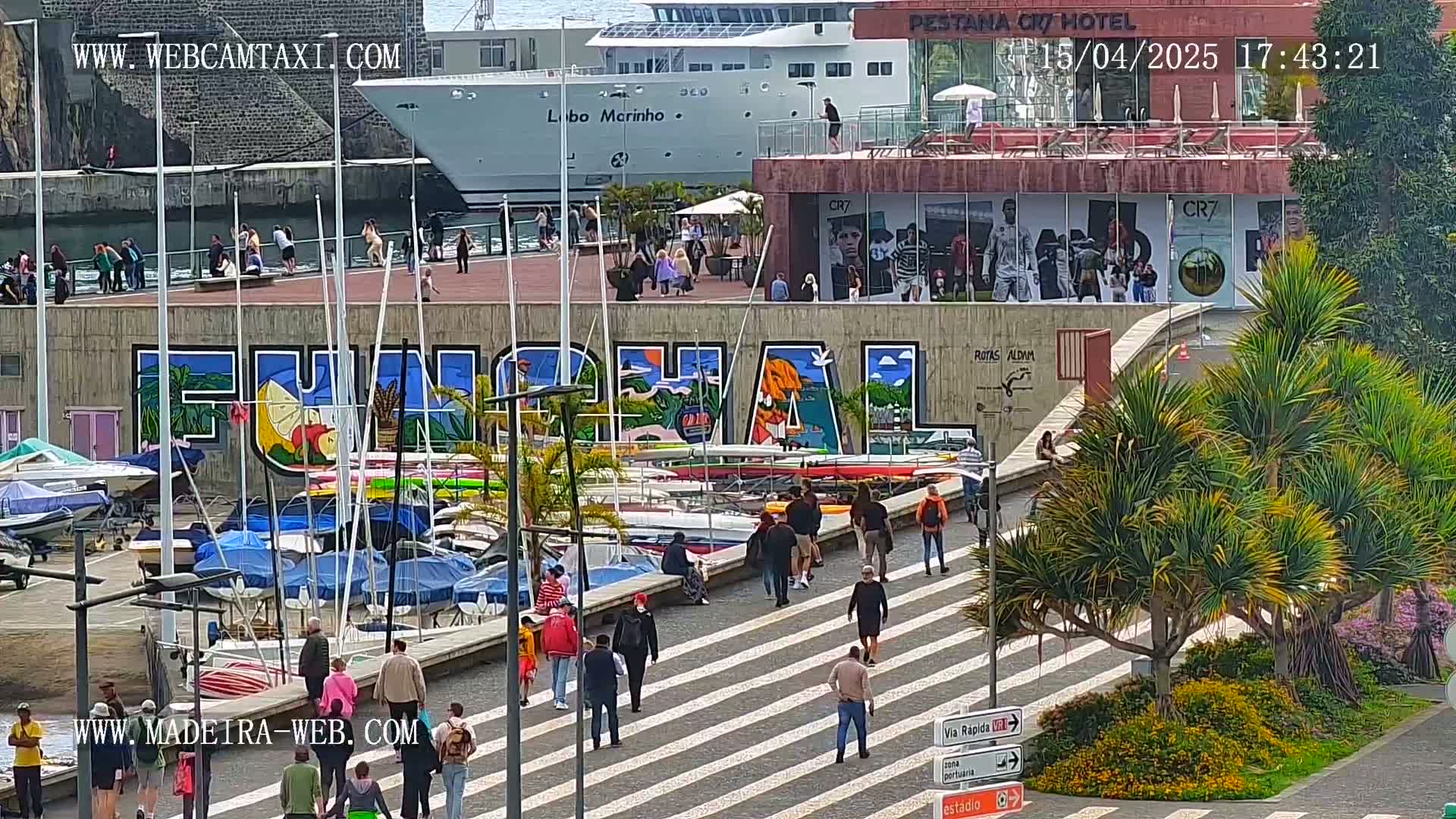A harbor scene on a sunny day shows a large mural of the word "Funchal" on a wall next to a marina filled with boats, with people walking along a pedestrian crossing in the foreground and a large cruise ship in the background.