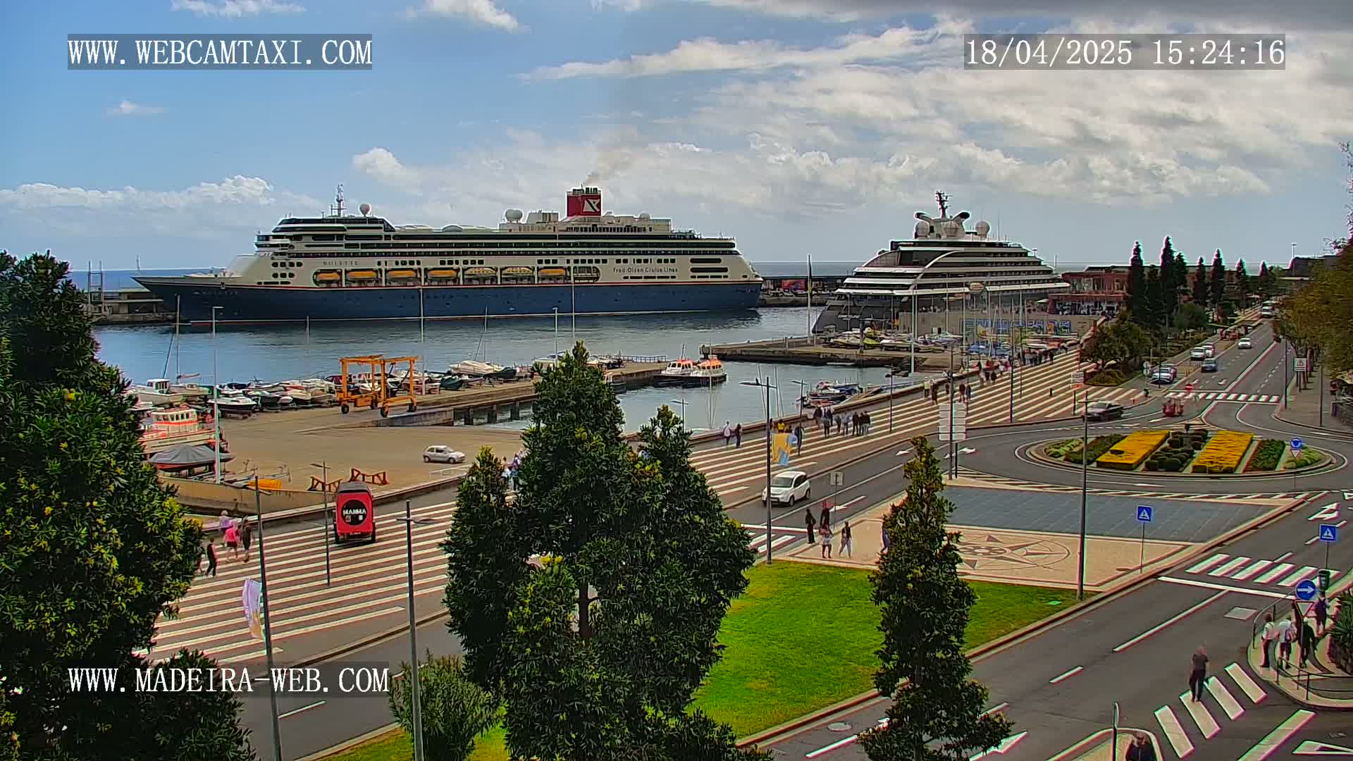 Two large cruise ships are docked at a harbor on a sunny day, with a paved area and pedestrian walkway leading to a roadway with cars and people.