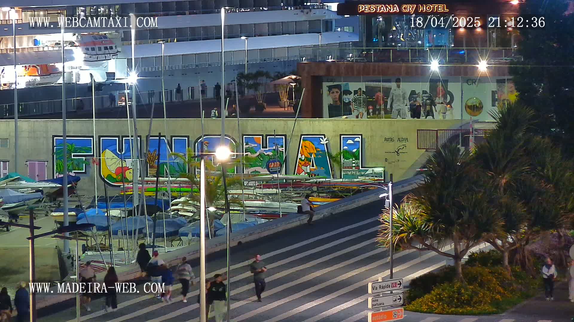 At night, a harbor scene shows numerous small boats docked along a wall with large, colorful graffiti of the word "Funchal," pedestrians walking on a nearby crosswalk, and a large building with images of people on its facade, all under nighttime lighting.