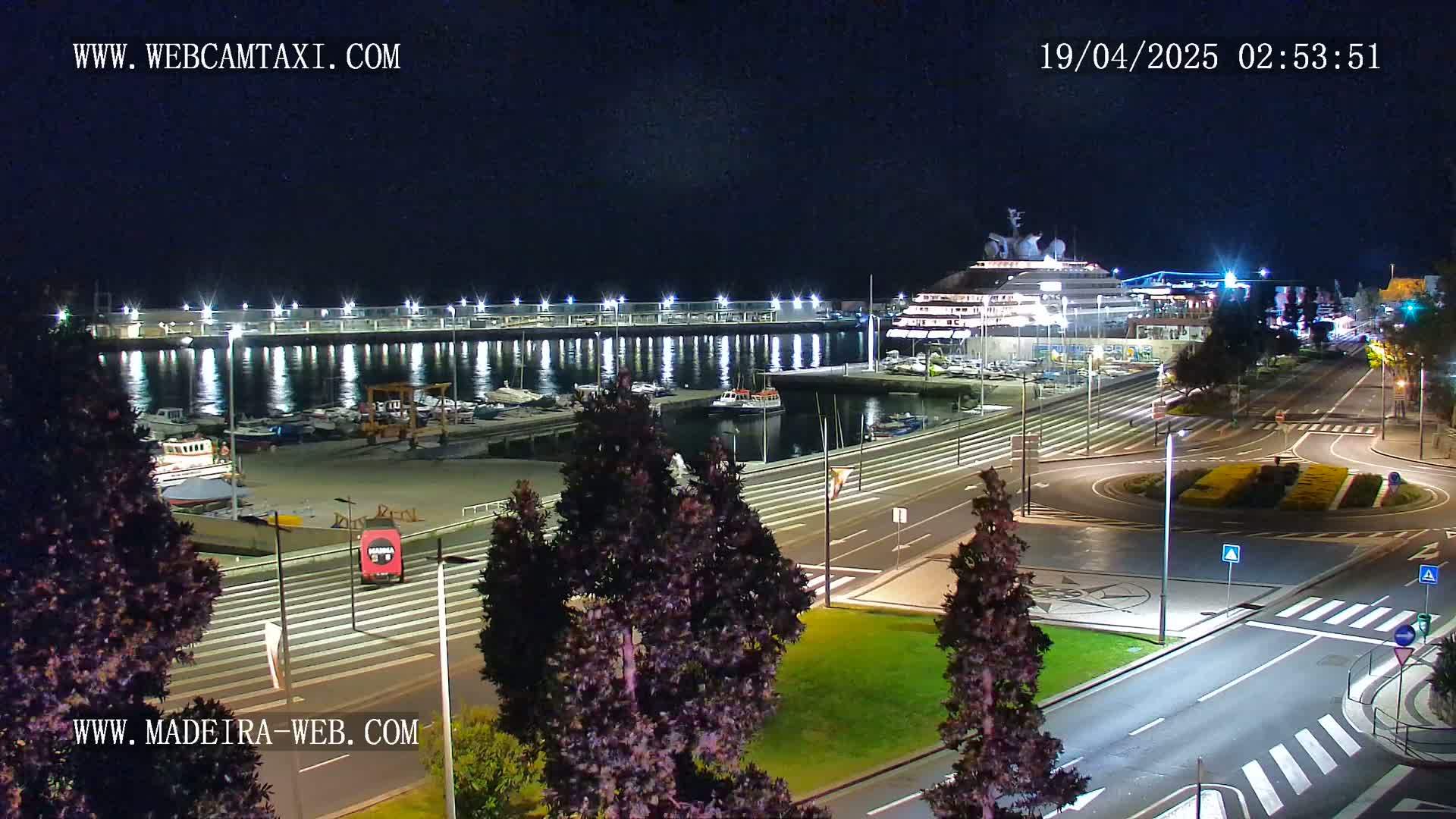 A large yacht is docked at a marina at night, with calm water reflecting lights on the shore and surrounding streets are mostly empty.