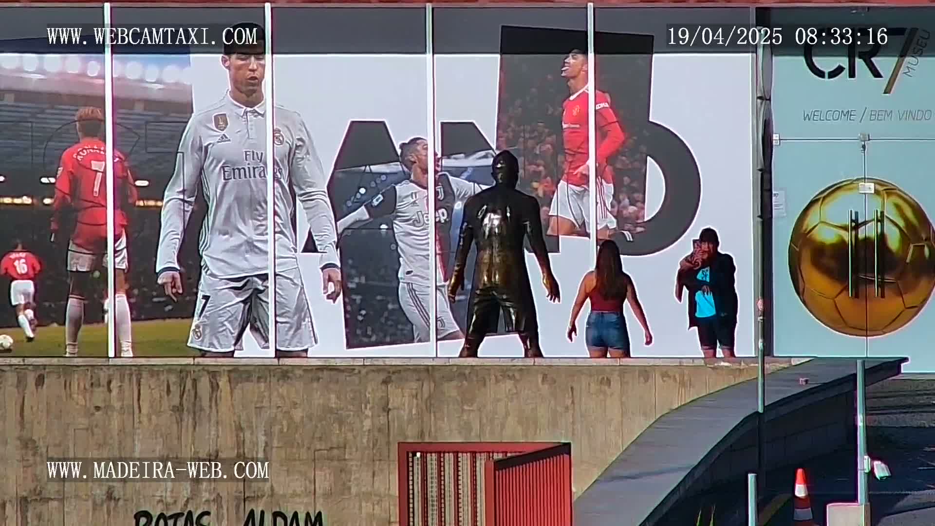 A large outdoor billboard displays images of Cristiano Ronaldo in various soccer uniforms, along with a bronze statue of him, while two people stand in front of it on a sunny day.