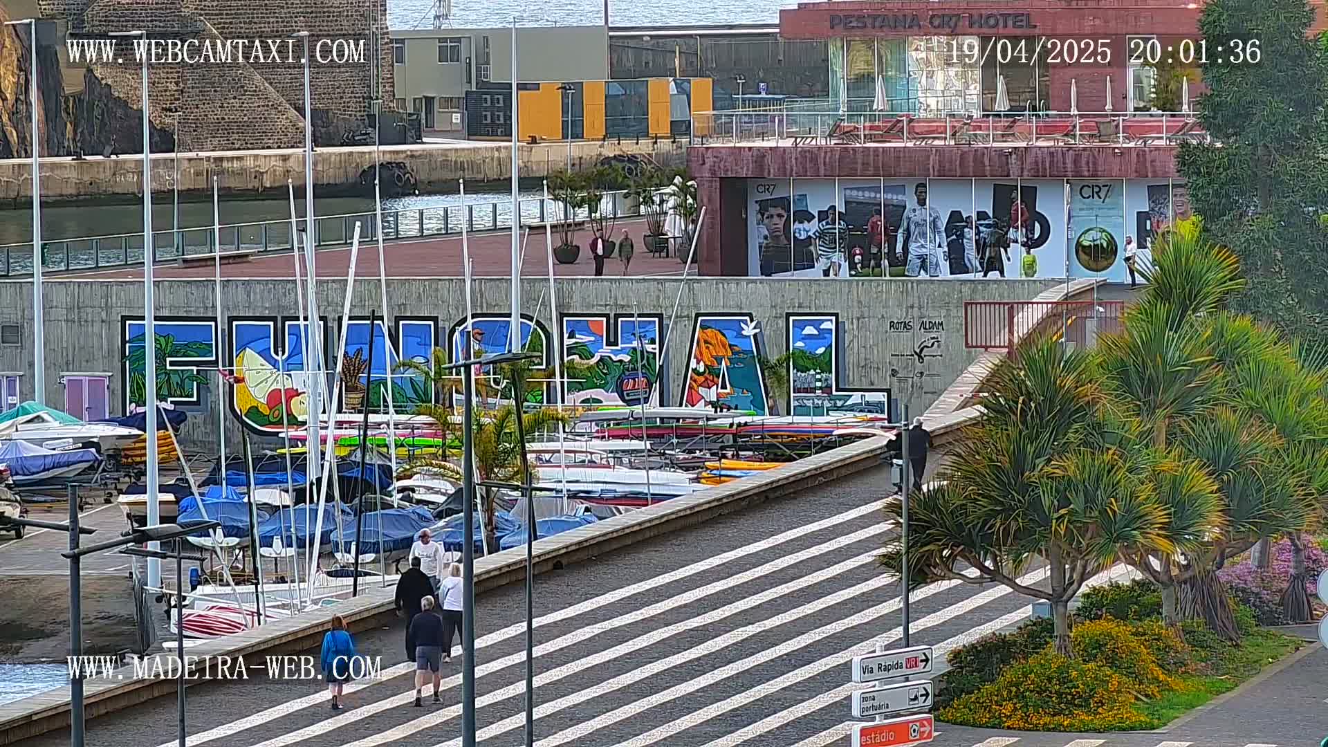 A marina filled with sailboats and small motorboats sits below a mural that spells out "Funchal," with several people walking along a striped walkway and palm trees in the foreground, under a partly cloudy sky.