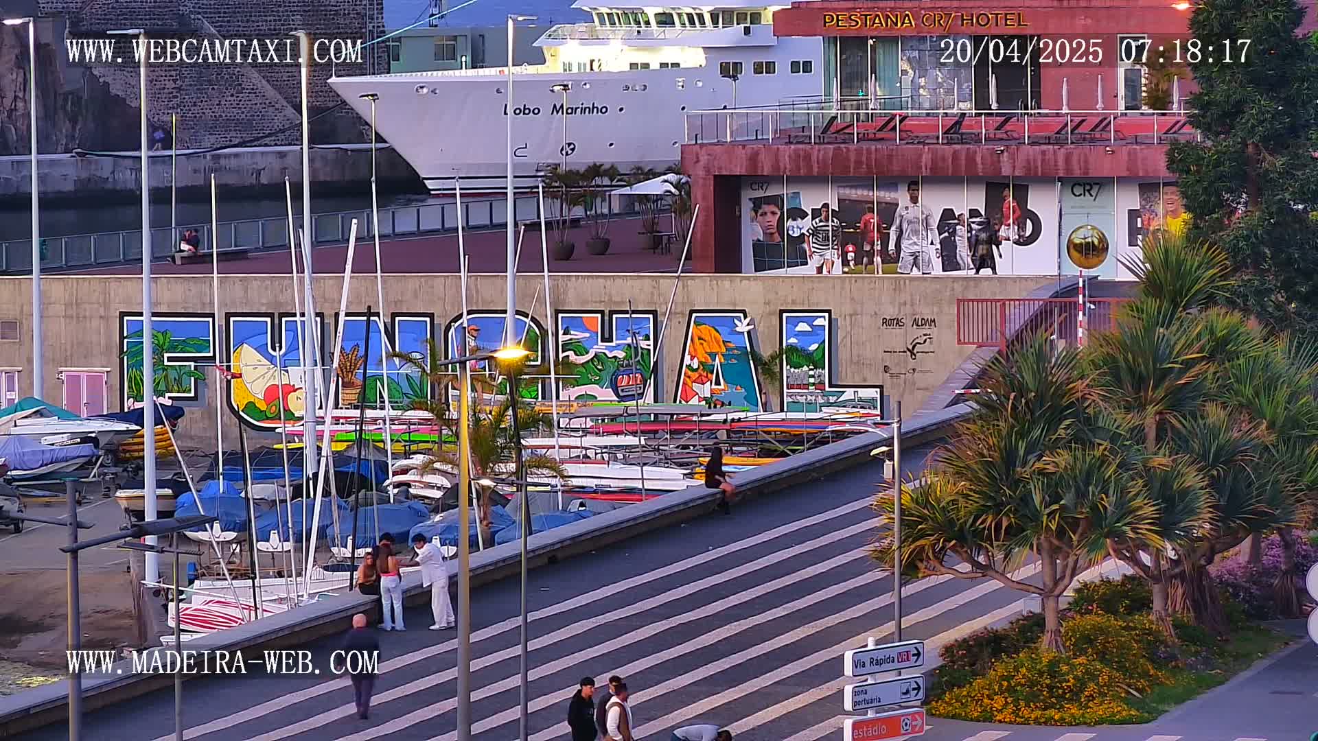 A harbor scene at dusk shows a large mural of "Funchal" on a building, numerous boats docked, a cruise ship, and palm trees along a walkway.